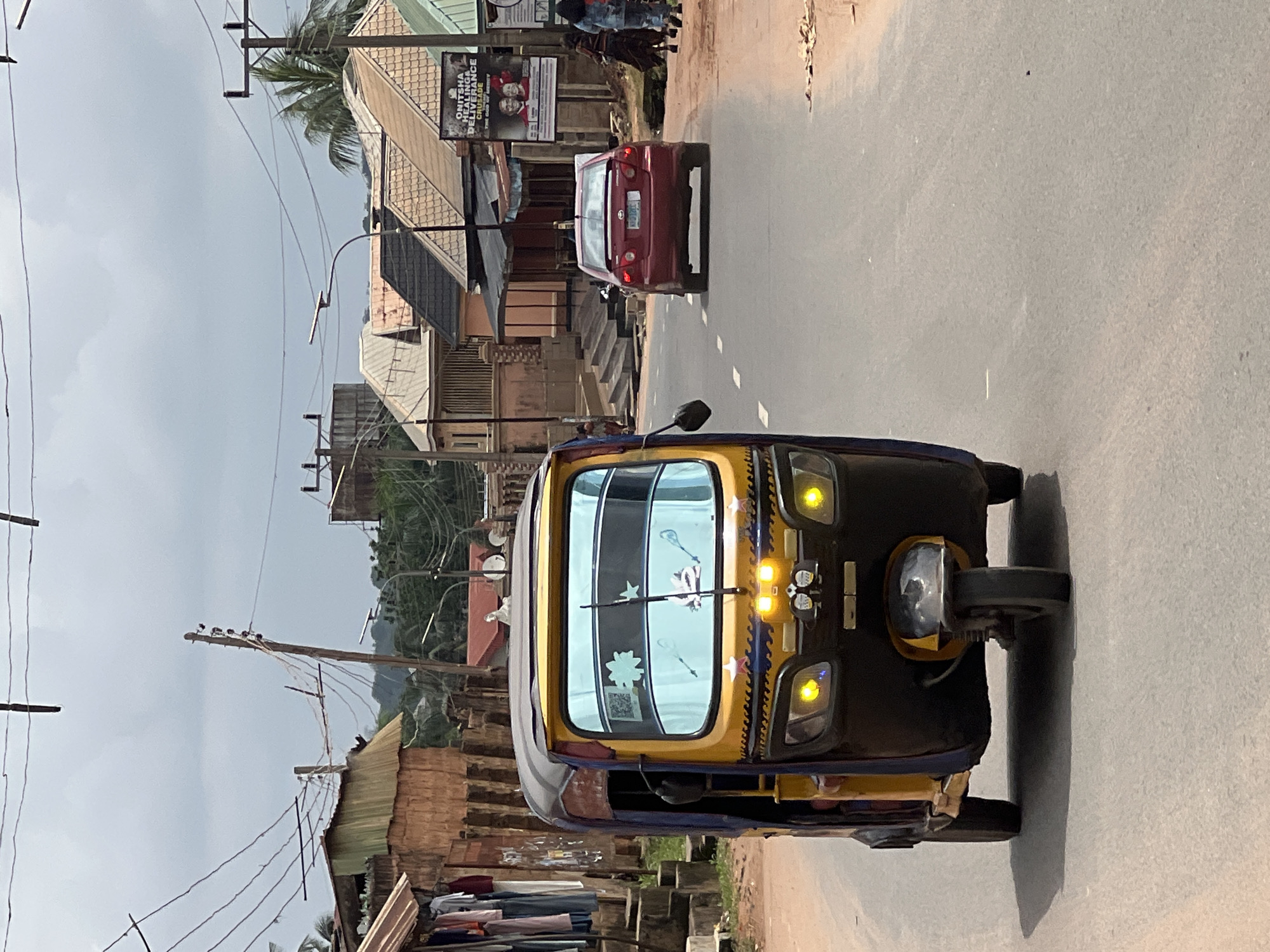 In Nkpor, Nigeria, a yellow and blue auto-rickshaw drives down a paved road under an overcast sky. The auto-rickshaw has a blue tinted windshield with white star decals and two windshield wipers. Two yellow headlights are illuminated on the front of the vehicle. To the right of the auto-rickshaw, a red sedan is also driving down the road. On the right side of the road, several buildings are visible, including shops with clothes hanging outside and a large poster advertising a church event. A few people are walking on the sidewalk next to the buildings. Utility poles and power lines are visible in the background. The road is mostly clear, with some debris scattered on the right shoulder.