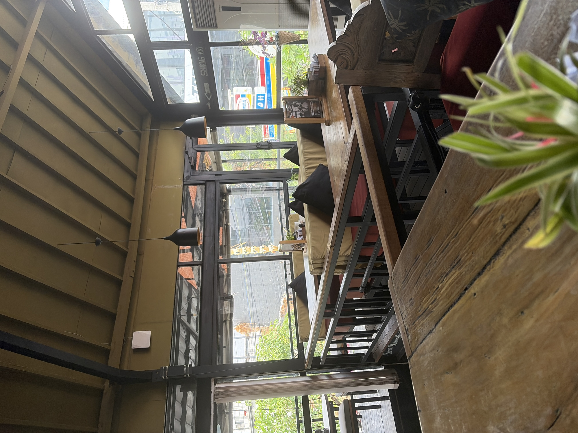 The image shows the interior of a restaurant in North Kuta, Indonesia, with a focus on the dining area and a view of the street through large windows. In the foreground, a rustic wooden table occupies the lower portion of the frame, with a blurred plant in the bottom right corner. Beyond this table, several dining tables are set with chairs and cushions, arranged on a raised platform. The tables are made of wood, and the chairs appear to have wooden frames with fabric upholstery. Black throw pillows are placed on the benches.
The restaurant features an open-air or semi-open-air design, with a sloped roof made of exposed wooden beams. Large glass windows and sliding doors line the perimeter, offering a view of the outside. Through the windows, the urban landscape of North Kuta is visible, including buildings, traffic, and greenery. A "SMOKING ARE" sign is visible on the window. The overall ambiance suggests a casual, tropical dining setting. There are no people clearly visible in the image.