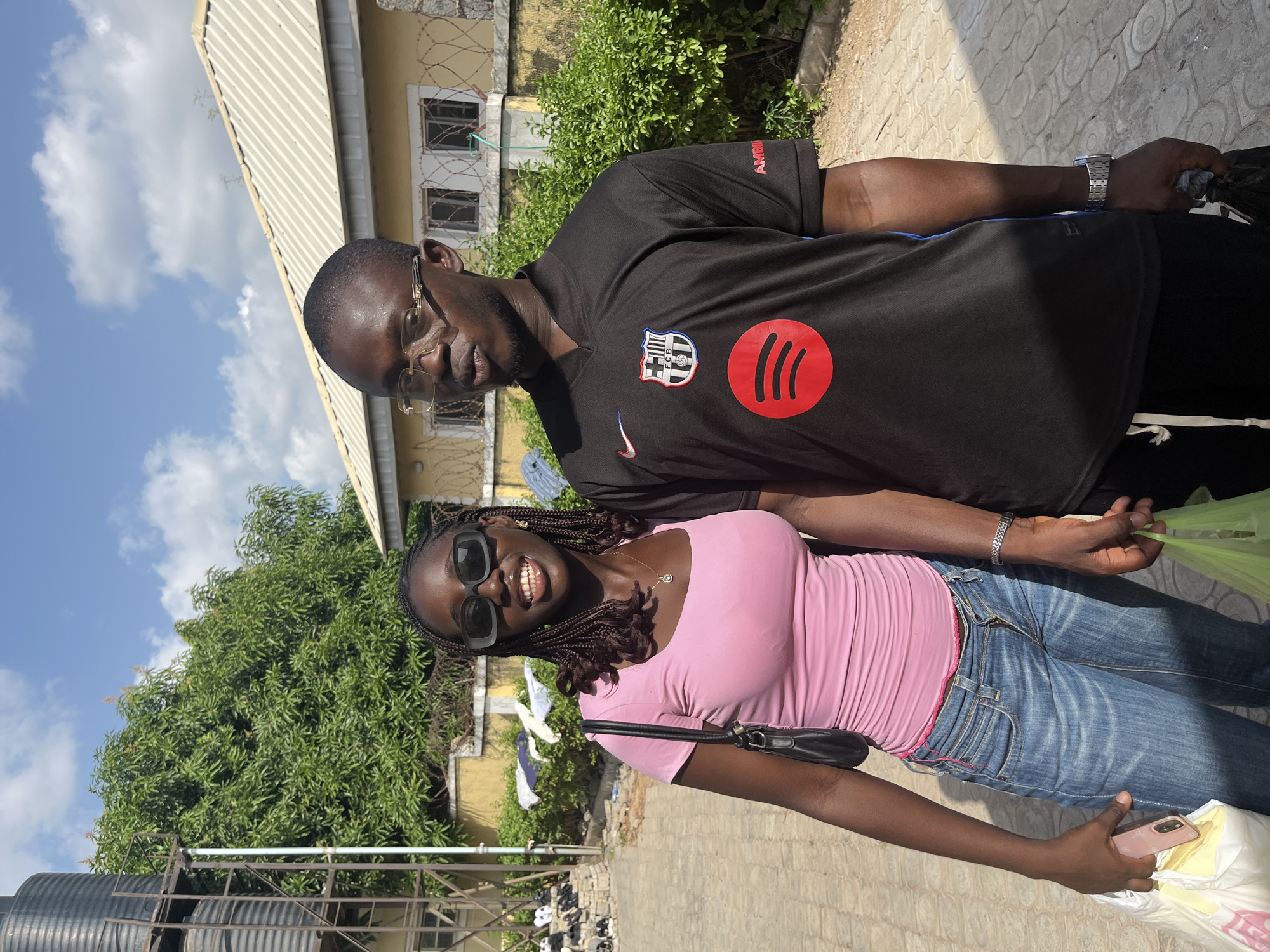 A photograph shows a man and a woman outdoors, likely in Abuja, Nigeria. The man, who is older, has glasses and wears a black soccer jersey with Barcelona’s crest and a Spotify logo. He has a watch on his left wrist. The woman, who is younger, wears a pink shirt, jeans, and sunglasses. She carries a small black purse. The two appear to be smiling and posing for the photo. In the background, a building with a tan exterior is seen, partially obscured by trees. Additionally, a water tank is visible. The sky is bright blue with few clouds. The location seems to be a residential or commercial area in the city.