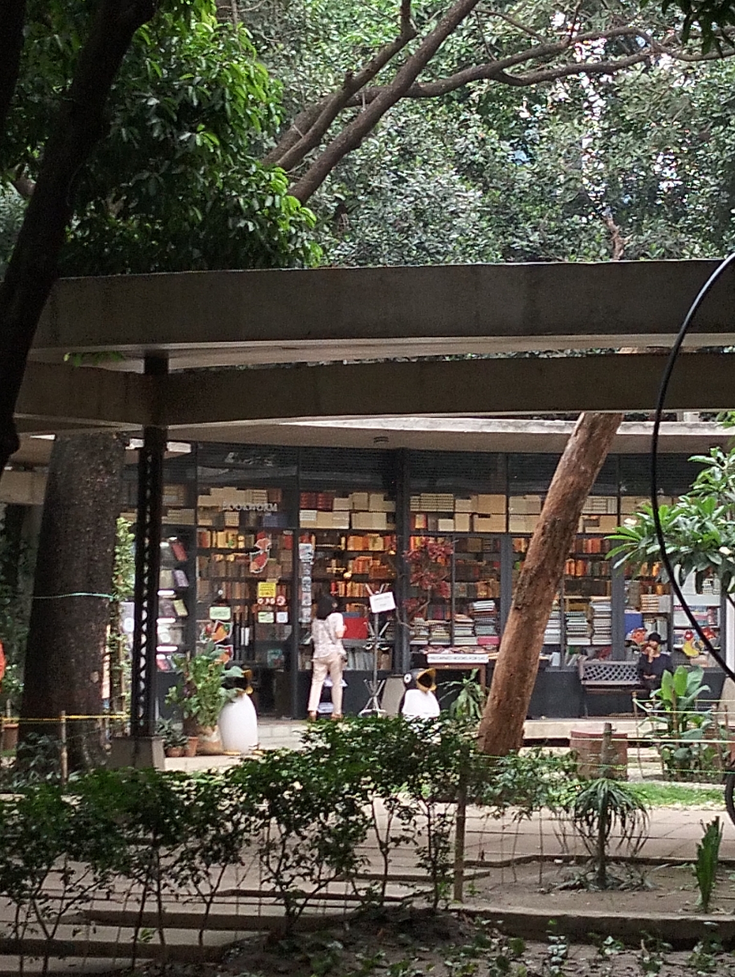 A mid-level shot depicts an outdoor scene with a bookshop in the backdrop. The location is in Dhaka, Bangladesh. The bookshop has glass windows revealing shelves filled with books. Two people stand in front of the bookshop, one with their back to the camera. A person sits on a bench towards the right side of the frame. In the foreground, there is foliage, with a concrete structure over the shop. Trees surround the structure.