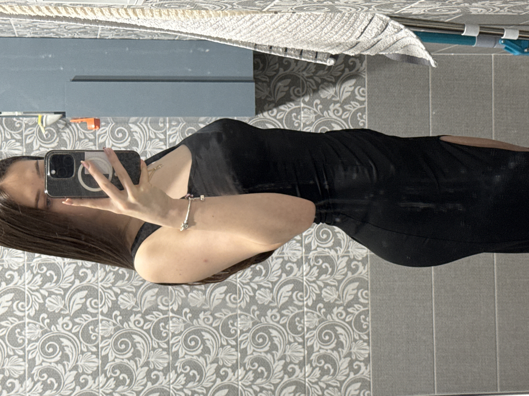 A person is taking a mirror selfie in a bathroom. The person has long, dark hair and is wearing a black dress. The bathroom has patterned wallpaper and grey tiles. A towel rack with a white towel is visible above the mirror. The location in Sofia, Bulgaria, cannot be determined from the image.