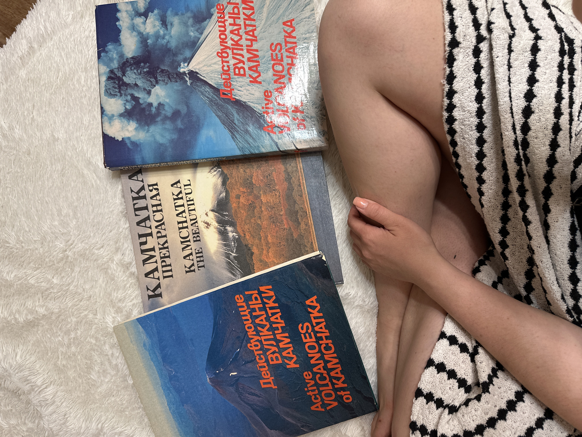 The image shows three books about Kamchatka, Russia, placed on a white, fluffy surface, likely a rug or blanket. The books are titled "Active Volcanoes of Kamchatka" (in both Russian and English) and "Kamchatka the Beautiful" (in both Russian and English). Adjacent to the books, on the right side of the image, are human legs and an arm covered in a white and black patterned towel. The legs are bent. The hand is resting near the knee. There is no indication of Nizhny Novgorod.