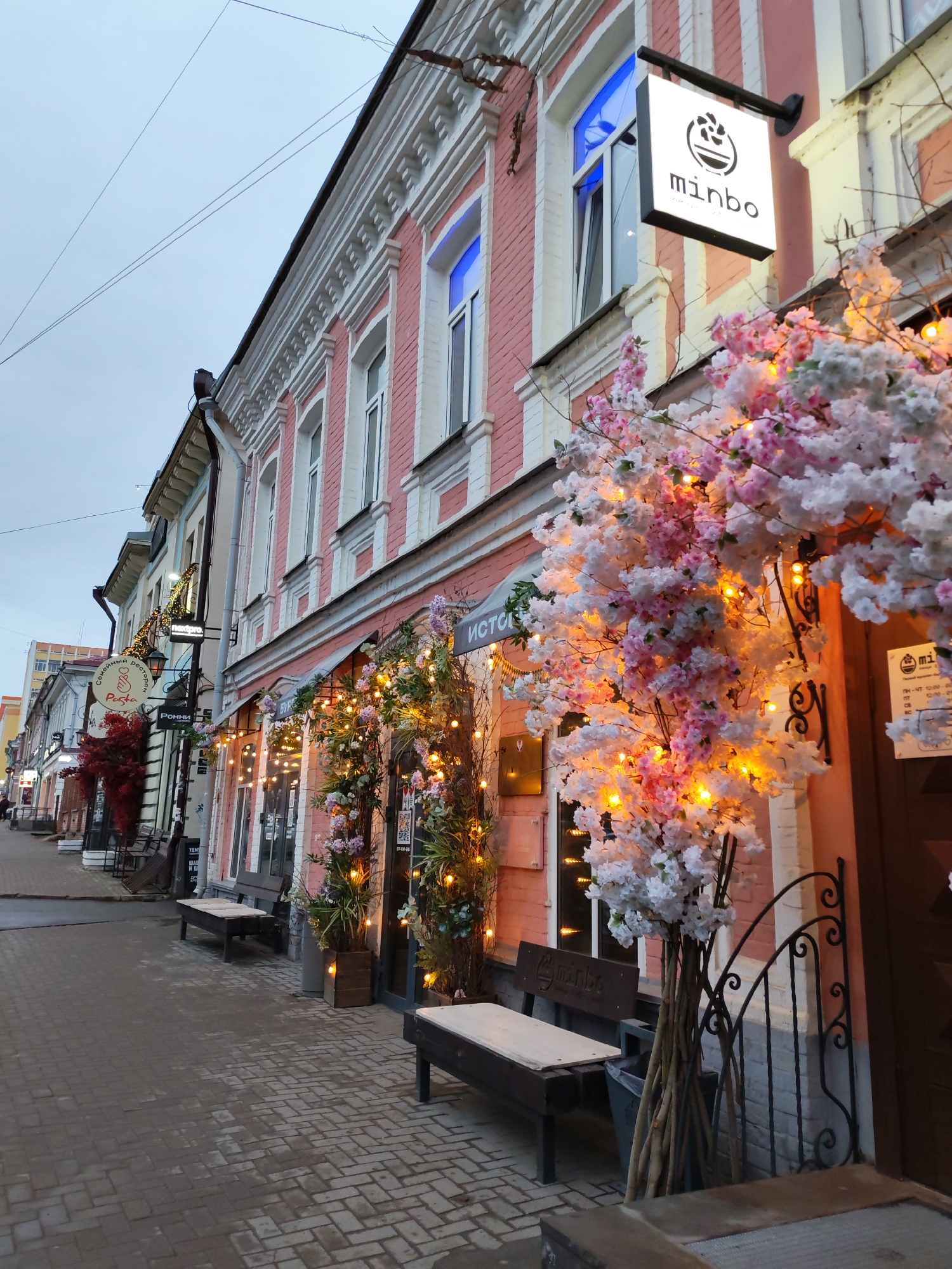 This is a street scene in Izhevsk, Russia. The street is paved with grey bricks and lined with buildings, mostly two or three stories high. The buildings are decorated for nighttime with lights, greenery, and flowers. The lighting consists of string lights and lights embedded in the flower and greenery decor. The building closest to the viewer is pink with white trim. It is the "minbo" restaurant. Near the storefront are benches. The sky is overcast. Several other businesses are visible, including a pasta restaurant called "Pasta Modok". The scene is peaceful and inviting.