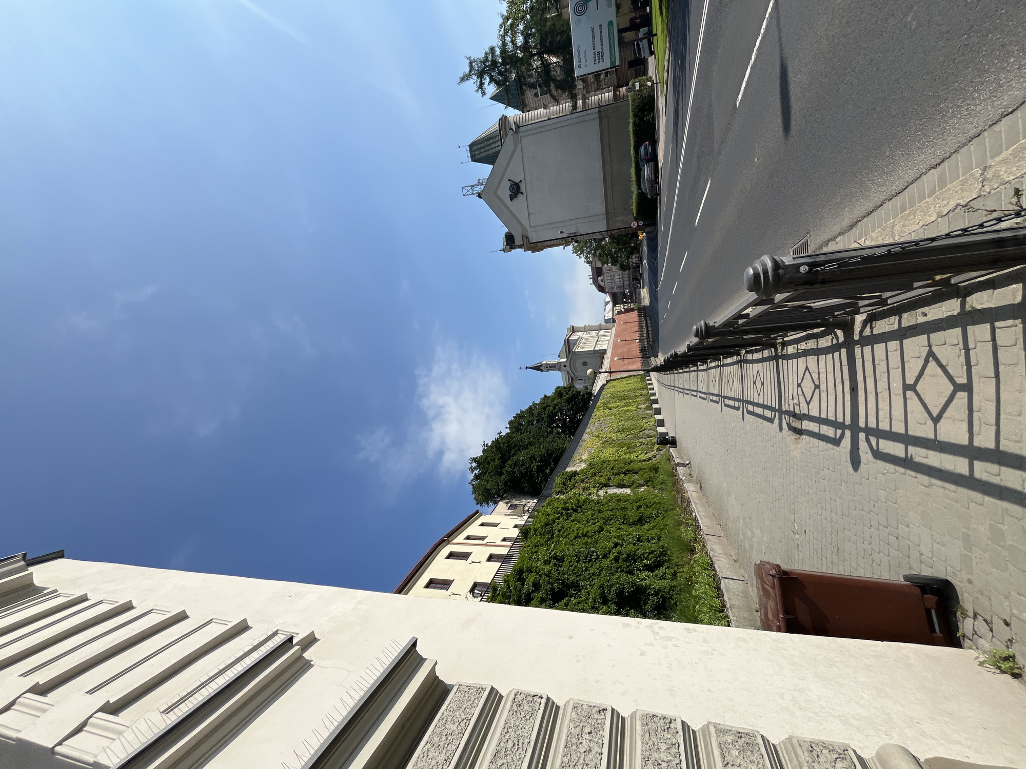 In an urban scene under a clear blue sky with scattered clouds, a wide asphalt road runs diagonally from the middle ground to the right. The road has double white lines dividing lanes and exhibits a slight sheen from the sunlight. A textured metal fence with decorative posts separates the road from a paved sidewalk on the left. This sidewalk is made of light-colored rectangular pavers and is partially in shadow cast by the fence. A brown wheeled trash bin is positioned on the sidewalk near the left edge of the frame. To the left of the sidewalk, a light-colored building with textured stone and smooth stucco sections rises upwards, its facade featuring linear architectural details and what appear to be decorative spikes along the top edge. Beyond the building, a long, high wall covered in dense green ivy stretches into the distance. Further down the street, more buildings are visible, including a structure with a tower and a spire, as well as another building with an ornate roof and a triangular gable. A large billboard advertising a company named "Zorbmotors" is also present. The environment suggests a sunny day in a city, possibly with historical architecture.