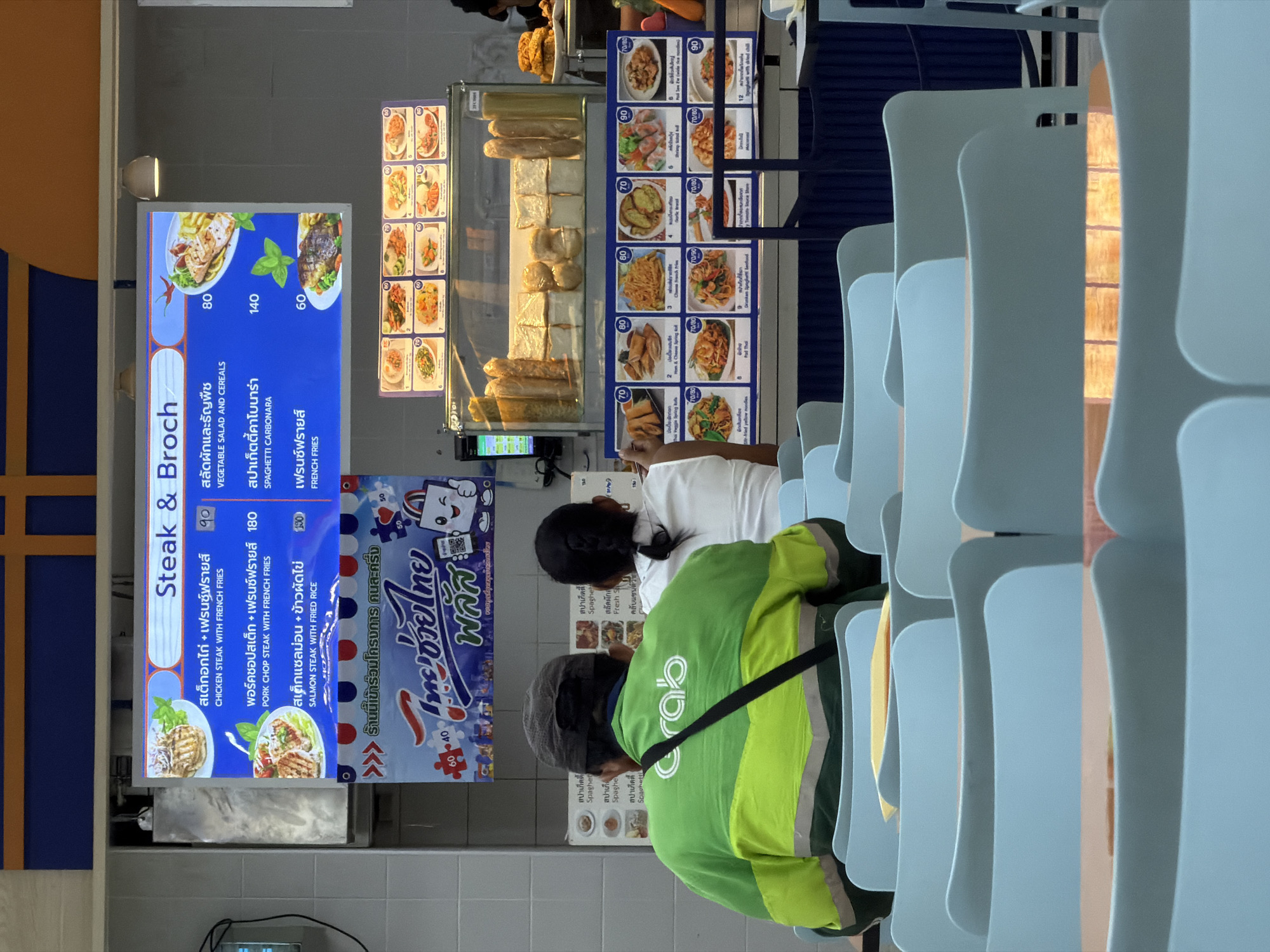 The image depicts a food court setting in what appears to be Thailand. Two individuals are visible, both facing away from the camera. One person is wearing a bright green uniform with a "Grab" logo, suggesting they are a delivery driver. The other person is wearing a white shirt.
In the foreground, a brightly lit menu board displays food items in Thai and English. The menu features "Steak & Broch," with options like "Chicken Steak with French Fries" (90 THB), "Pork Chop Steak with French Fries" (180 THB), and "Salmon Steak with Fried Rice" (190 THB). Other menu items like "Vegetable Salad and Cereals" (80 THB), "Spaghetti Carbonara" (140 THB), and "French Fries" (60 THB) are also listed.
To the right of the menu board, a display case shows various food items, including bread, pastries, and what appear to be prepared meals, with prices listed. Another blue sign with Thai text and a cartoon character is visible, possibly advertising a promotion. The overall scene suggests a busy food establishment, likely during daytime hours, given the lighting. The presence of the "Grab" logo indicates food delivery services are available. The environment appears to be indoors, with rows of light blue chairs in the background.