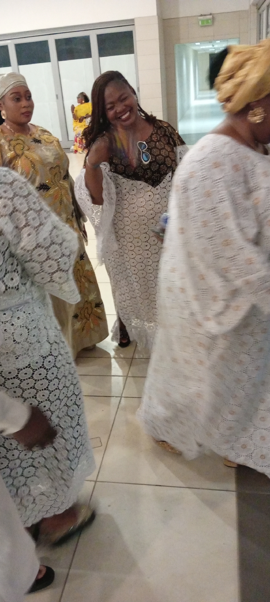 In the city of Gandu Albasa, Nigeria, a group of women are gathered indoors, likely for a celebratory event. The women are adorned in traditional African attire, characterized by vibrant colors and intricate designs. One woman, in the foreground, wears a flowing white lace gown with delicate circular patterns. She is laughing heartily, her head tilted back, and her mouth open. She wears sunglasses perched on her head. To her left, another woman is seen in profile, wearing a gold and white patterned outfit and a matching headscarf. Her expression is neutral, and she appears to be looking towards the left. In the background, a third woman in a bright yellow outfit is visible, her back to the camera, her arms raised. The floor is tiled with a light-colored material, and the lighting suggests an indoor environment.