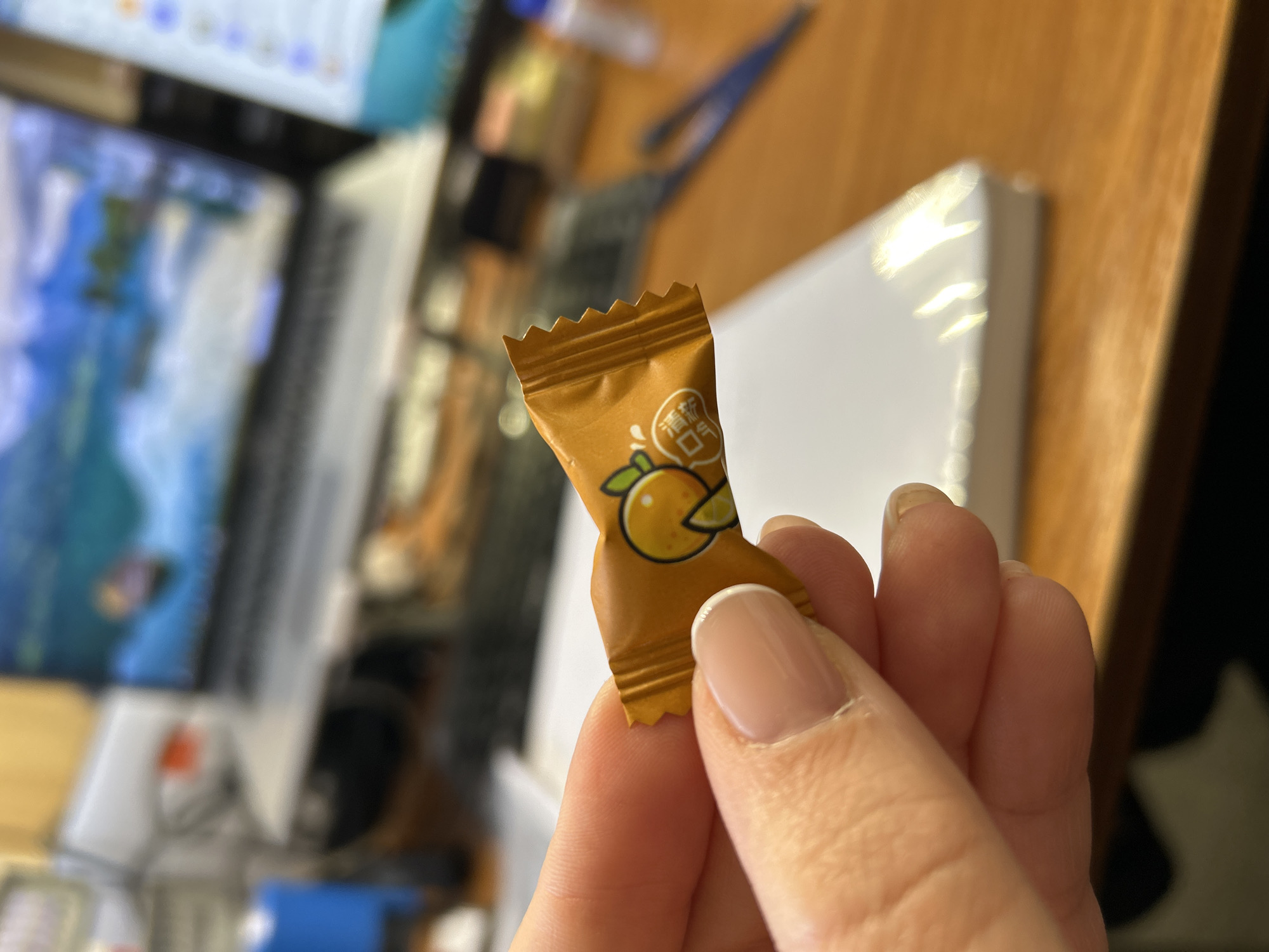 A person's hand holds an opened piece of orange candy, adorned with a drawing of an orange and Chinese lettering. The candy wrapper is a light brown color. The hand has well-manicured nails. The background reveals a cluttered desk, likely in an indoor setting. A computer monitor sits to the left, displaying indiscernible content. Miscellaneous items, including a white box, are scattered around the desk. The scene does not appear to be in Ural'skoe, Russia.