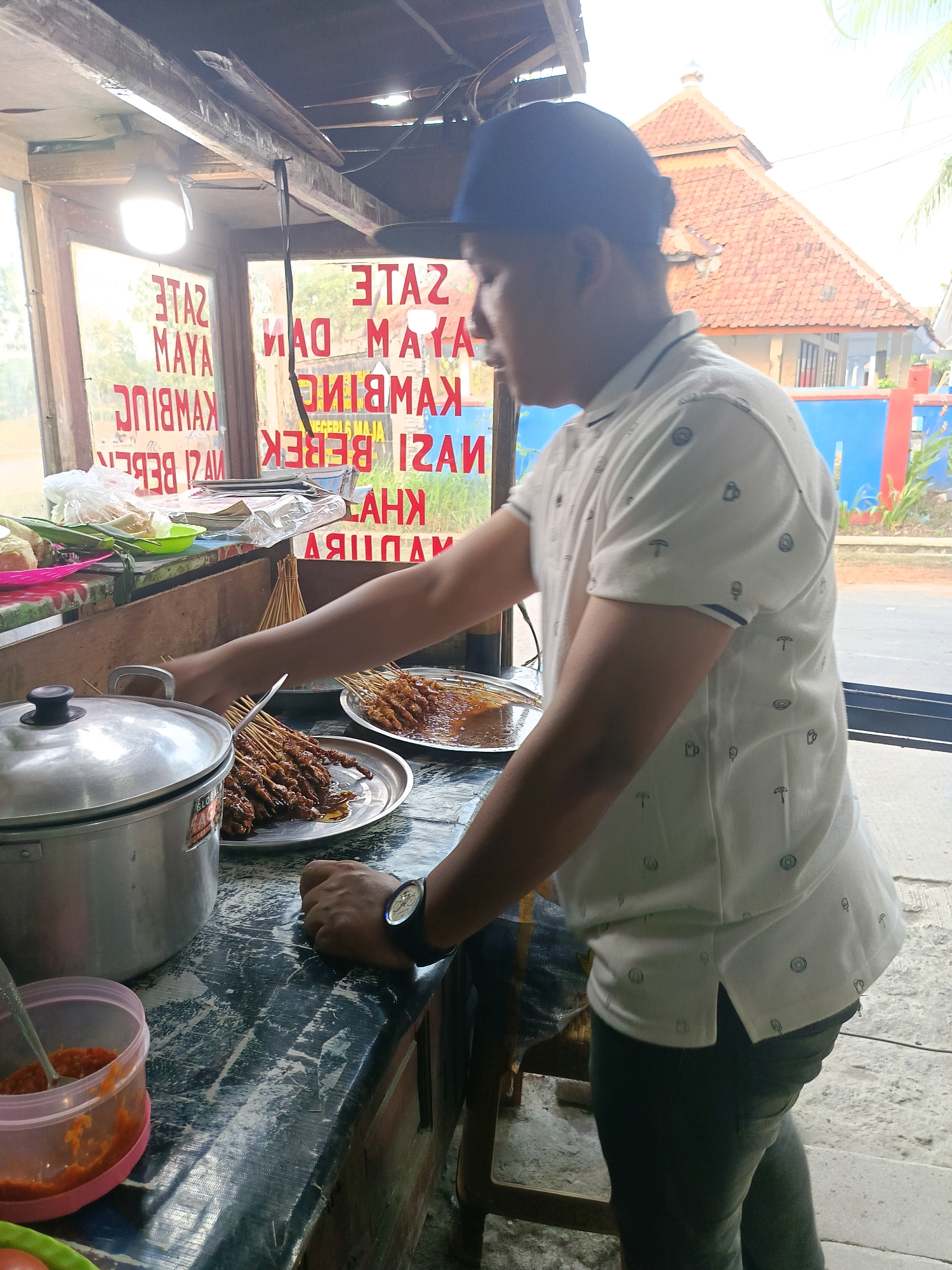 The image depicts a man in a white, patterned polo shirt and a dark baseball cap, standing in what appears to be a food stall in Pasir Kembang, Indonesia. He is shown reaching towards a plate of satay, with skewers of grilled meat arranged on it. In the foreground, a large silver pot is visible on the left, next to a small pink container holding red chili sauce. The background shows a window with signage, partially obscured by the man, listing various food items such as "Sate Ayam," "Nasi Bebek," and "Kambing." Beyond the window, an outdoor scene is visible, including a building with an orange tiled roof and greenery. The lighting suggests it is daytime. The stall appears to be constructed of wood and glass.