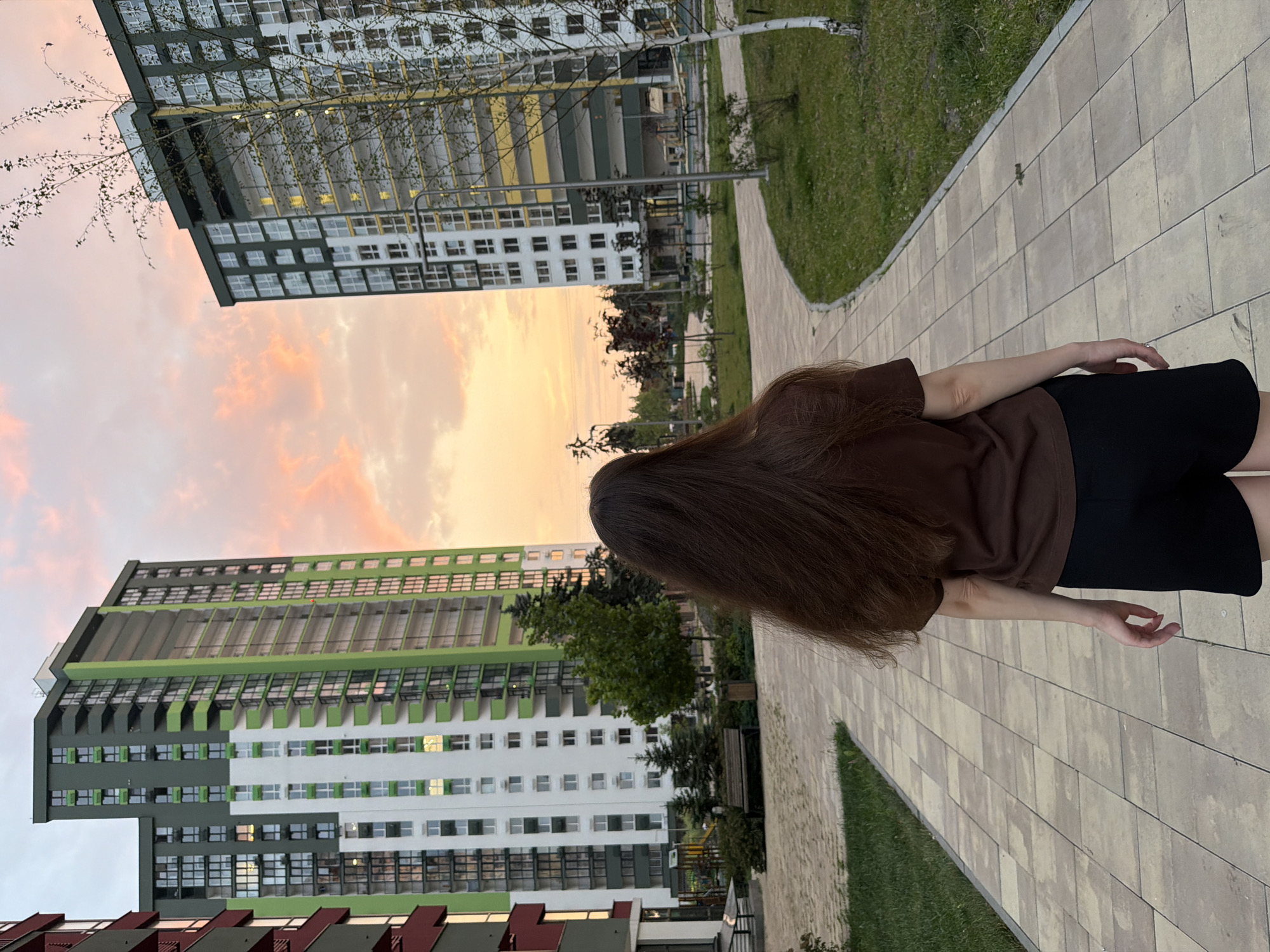 An adult female with long, dark brown hair is shown from behind, walking away from the camera on a paved pathway. She is wearing a brown t-shirt and black shorts. The pathway is flanked by green grass. In the background, two modern, multi-story apartment buildings are visible. The building on the left is primarily white and green, with a section of dark red. The building on the right is white and dark green, with yellow accents. The sky above displays a soft sunset with orange and pink clouds. The overall scene suggests a residential area within a city, possibly Kyiv, Ukraine, given the architectural style and the lighting conditions indicative of dusk.