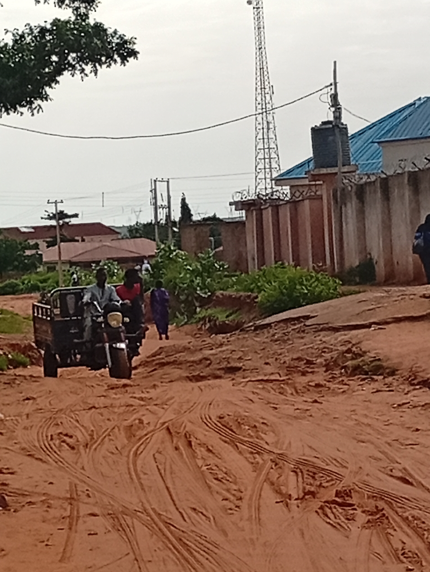 In Tumfure, Nigeria, a dusty, unpaved road dominates the foreground, marked by tire tracks and reddish-brown soil. A three-wheeled vehicle, resembling a motorcycle with a cargo bed, is the central focus. Two individuals are seated on the vehicle; the driver, a man wearing a light-colored traditional garment, is on the left, and a passenger wearing a red shirt is behind him. To the right of the vehicle, a person in a purple garment walks away from the camera. In the background, lush green vegetation borders the road, and several buildings with reddish-brown roofs are visible. A tall telecommunications tower stands prominently against a pale sky, alongside other utility poles and wires. A concrete wall with a blue-roofed structure behind it extends across the middle ground. The scene captures a moment of daily life on a rural-urban fringe.