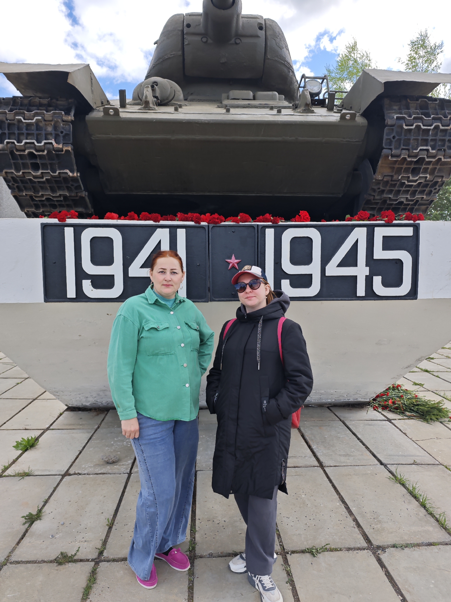 The image shows two women standing in front of a tank monument in Марковский, Russia. The monument has the years 1941 and 1945 displayed on it, separated by a star. Red flowers are placed along the base of the tank and on the ground to the right of the monument. The woman on the left is wearing a green shirt and blue jeans, and the woman on the right is wearing a black coat and a baseball cap. The ground is paved with concrete slabs. The sky is partly cloudy.