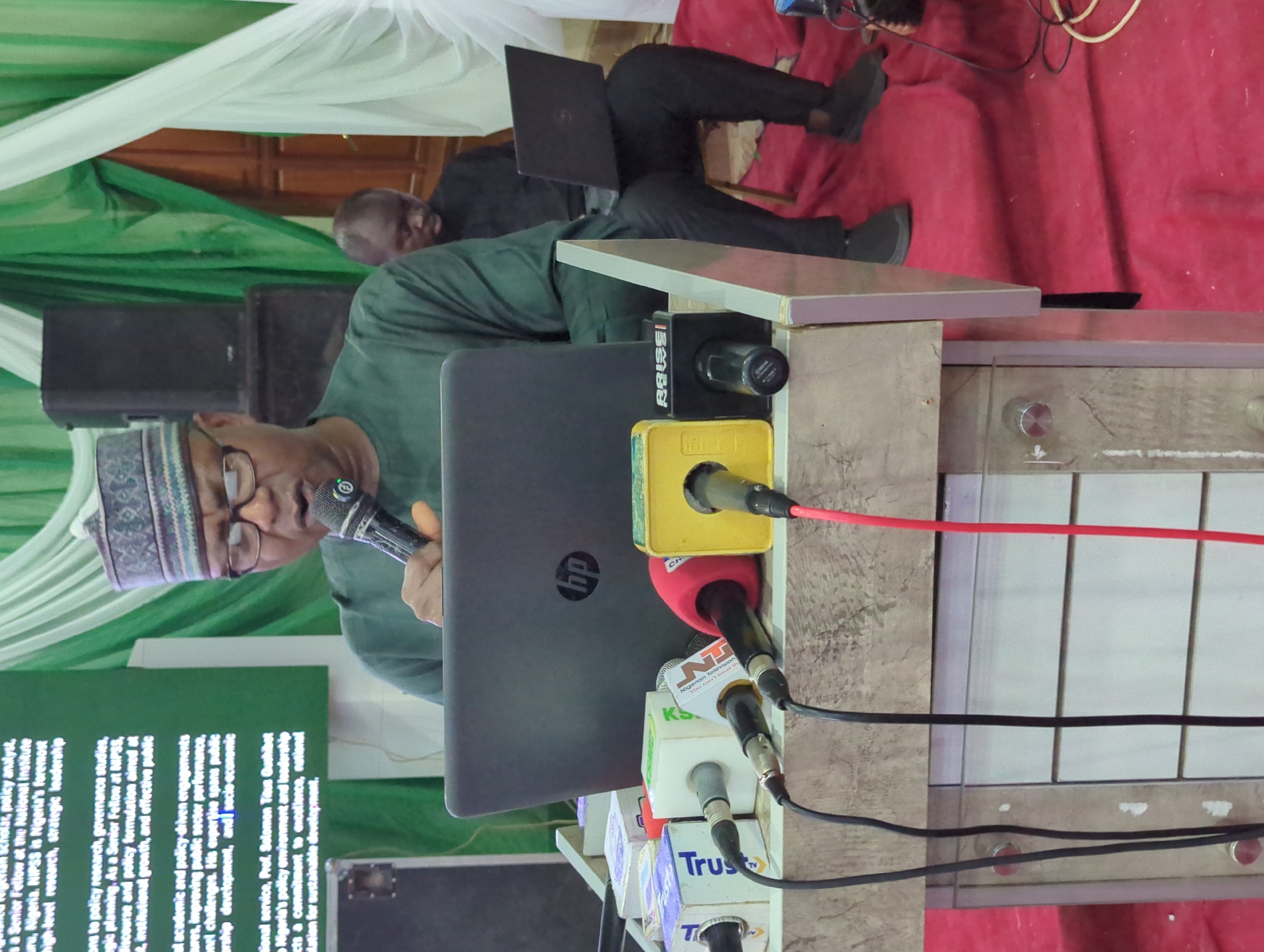 The image captures a man, presumably giving a speech, standing behind a podium with multiple microphones bearing logos of various news outlets, including "Nigerian Television," "Trust," and "Arise News." He is wearing a traditional cap and glasses, holding a microphone in his right hand, and facing a laptop with an HP logo. Another man sits in the background with a laptop on his lap. The podium is cluttered with wires and microphone attachments. A green and white draped cloth hangs in the background. The scene suggests a press conference or public event in Ungwan Liman, Nigeria.