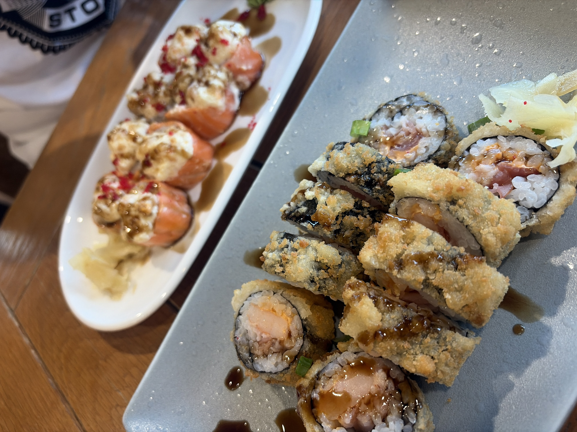 The image depicts two plates of sushi and sushi rolls arranged on a light brown wooden table in Saint Petersburg, Russia. In the foreground, a grey rectangular plate presents multiple deep-fried sushi rolls, predominantly featuring a crispy, golden-brown breaded exterior. Visible fillings include shrimp tempura and light pink fish, likely tuna or salmon, combined with rice and nori. These rolls are garnished with chopped green scallions and generously drizzled with a dark, glossy sauce, likely a sweet soy or eel sauce. A small pile of pale yellow pickled ginger is positioned on the right side of this plate. In the background, a white oval plate holds several pieces of salmon-wrapped sushi, characterized by bright orange salmon slices encasing rice and fillings, topped with a creamy white sauce and small red fish roe, also drizzled with the dark glossy sauce. A portion of pickled ginger is also visible on this plate. The wooden table displays a natural grain. A person's arm, wearing a dark garment with a visible patterned cuff showing the partial text "STO", is partially visible in the upper right background.