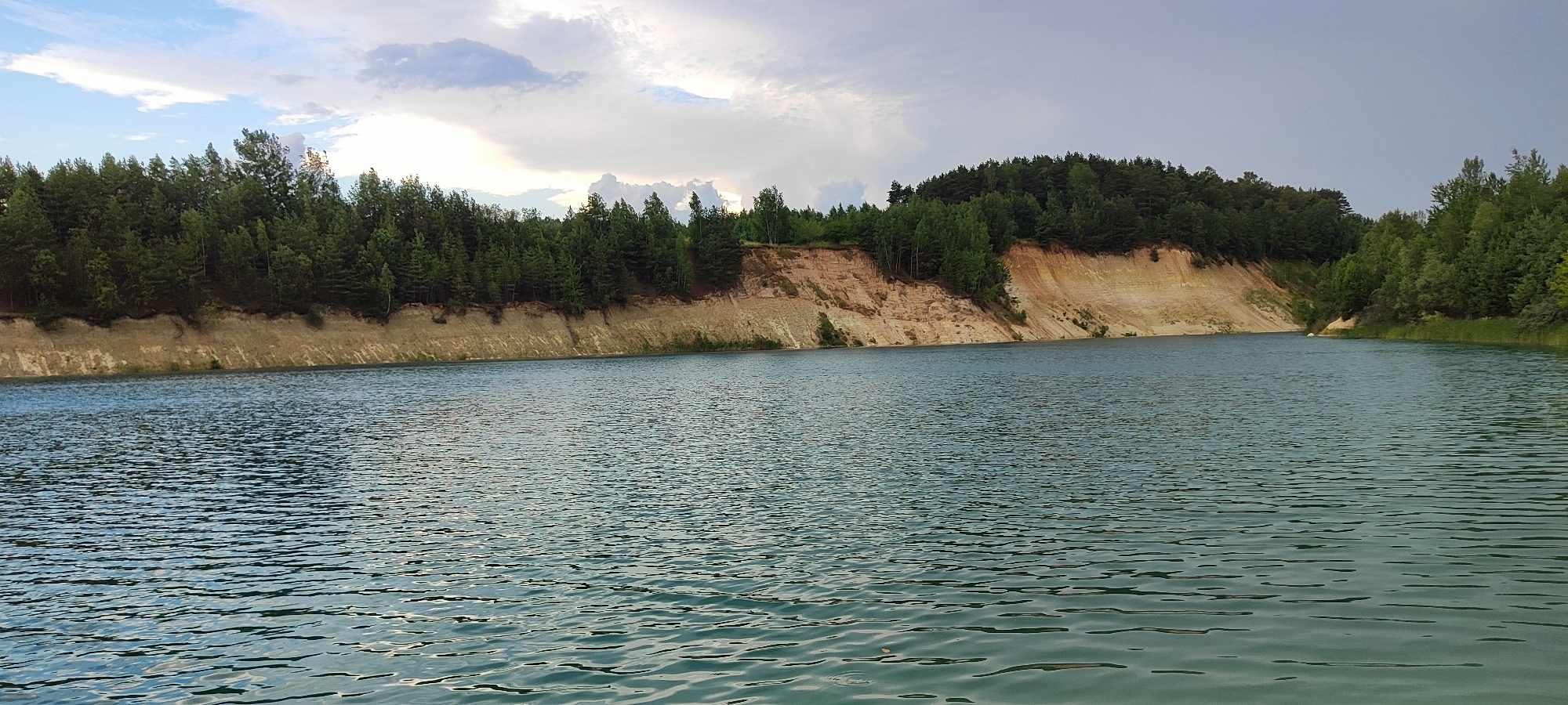 The image shows a panoramic view of a quarry lake with a steep, sandy embankment on the left side, topped with a dense forest of pine trees. The water is a clear, turquoise color with visible ripples on the surface. The sky above is partly cloudy, with patches of blue sky peeking through. The embankment shows distinct layers of sand and clay, with some vegetation growing at its base. The forest extends along the top of the embankment and continues to the right side of the image, creating a natural backdrop. The scene appears to be a tranquil and secluded natural landscape.