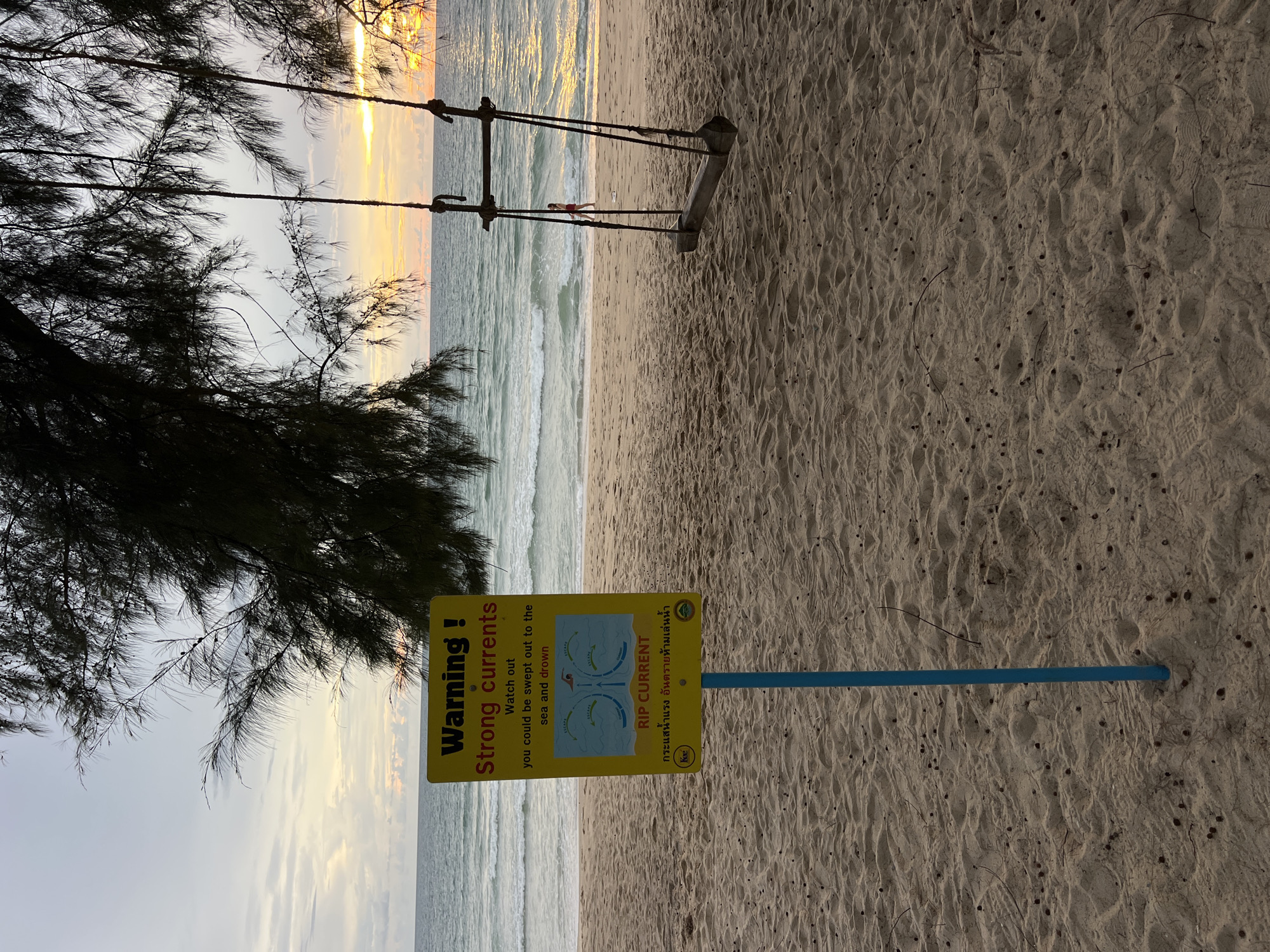 An outdoor, eye-level, medium shot captures a beach scene at dusk. The foreground is dominated by sand, with a weathered wooden swing set on the right, its ropes partially visible. To the left, a yellow warning sign stands on a blue pole, displaying text and an illustration depicting rip currents. The middle ground features the ocean with gentle waves and a sandy shore. In the background, the sea meets the horizon under a cloudy sky with hints of sunset colors. A single person is visible on the beach near the water. The overall atmosphere is serene, with the encroaching twilight casting a muted glow on the scene.