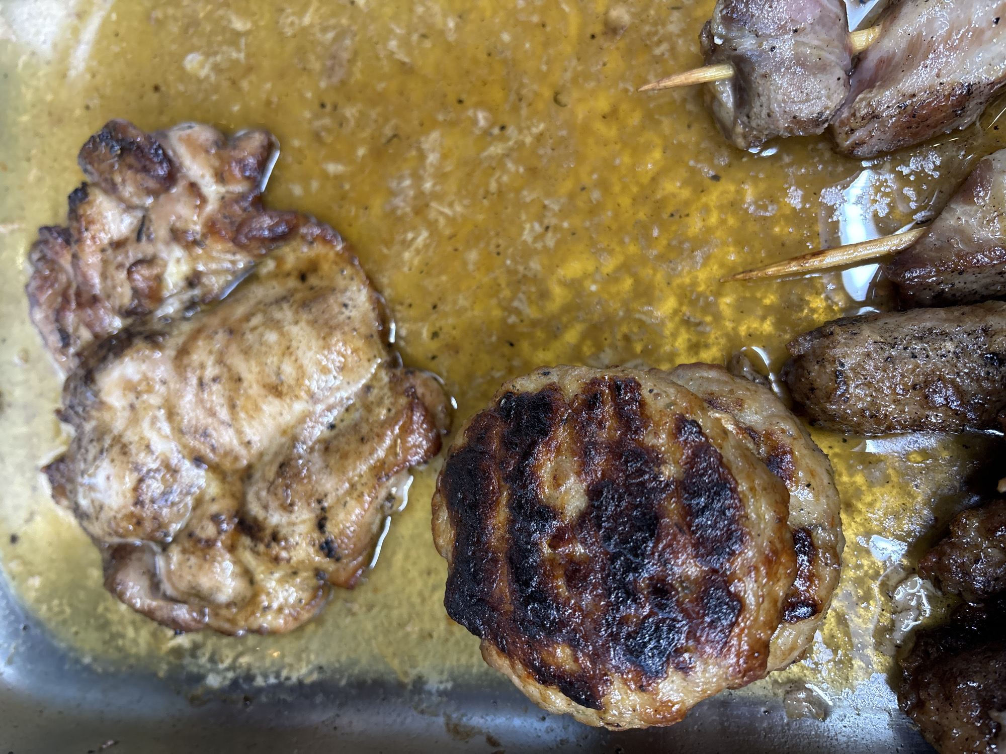 The image shows cooked meat in a pan filled with a yellow liquid, likely cooking oil or sauce. On the left, there's a piece of chicken with grill marks. To its right is a burger patty, darker in color, indicating it is more charred. At the top right corner, meat kebabs on wooden skewers are visible. The pan appears to be metal. There are no people visible in the image. The context of Sofia, Bulgaria is not evident in the image.