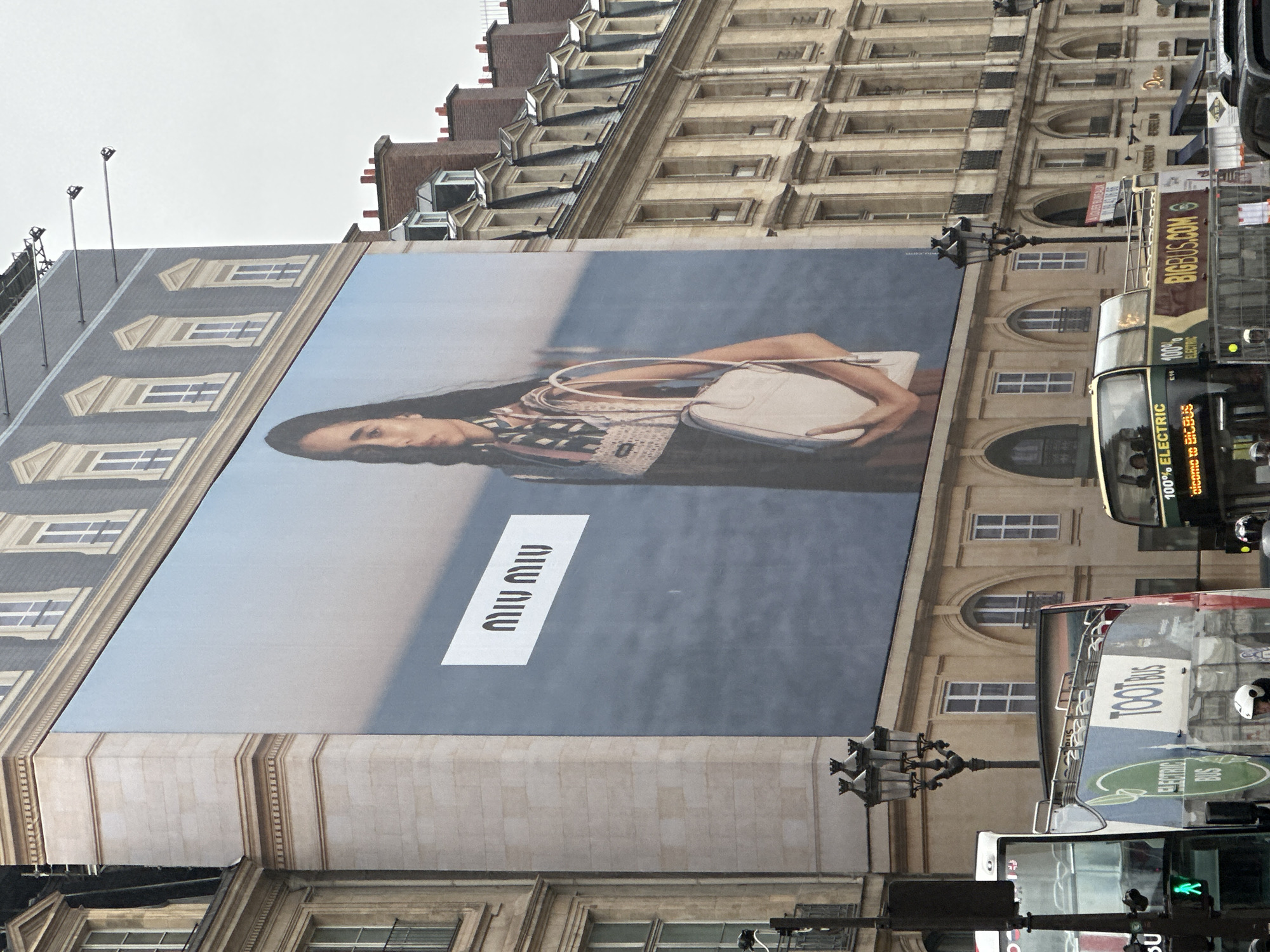 The image depicts a large advertisement for Miu Miu, a fashion brand, displayed on the side of a building in Paris, France. The building has multiple windows and architectural details typical of Parisian buildings. The advertisement features a woman with a Miu Miu handbag. In the foreground, there is traffic, including a double-decker bus labeled "TOOTBUS," which is marked as an electric bus, and another double-decker bus labeled "BIGBUS.COM." The weather appears overcast, suggesting a cloudy day. The image is taken from a ground-level perspective, looking up at the building and the advertisement.