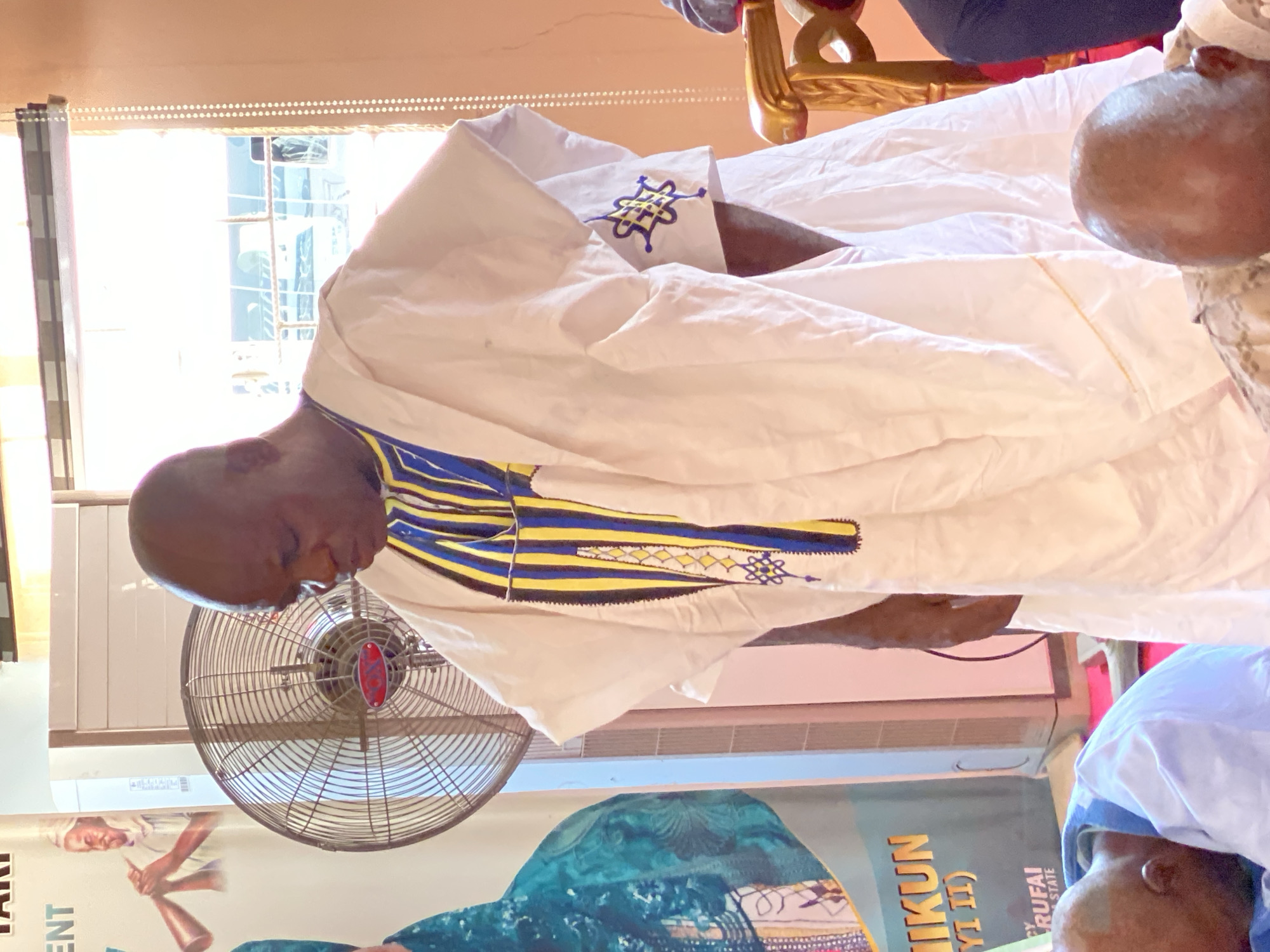 The image shows an indoor scene, likely in Sabon Tasha, Nigeria. A bald man in traditional white attire with blue and yellow embroidery is in the foreground. A fan stands nearby. A poster with an image of a person holding an object is also visible. Another person is partially visible in the foreground. The environment suggests a formal or public gathering.