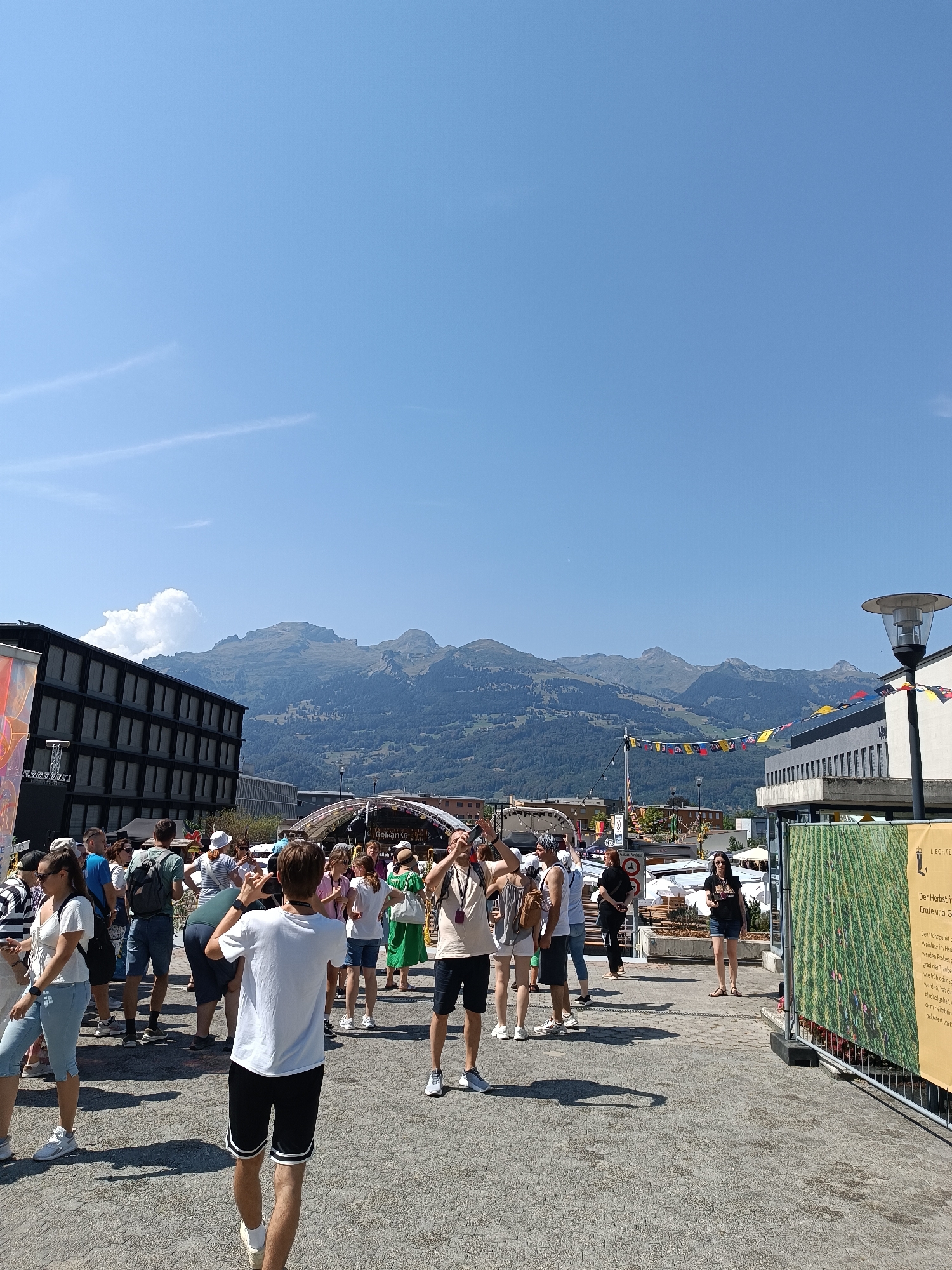 The image shows an outdoor scene in the city of Vaduz, Liechtenstein, on a sunny day. In the foreground, a crowd of people is gathered, many of whom appear to be tourists. Several individuals are holding up their phones, possibly taking pictures or videos. The ground is paved with cobblestones. Behind the crowd, there are buildings, including a modern dark-colored building on the left with many windows, and a lighter-colored building with a balcony on the right. In the background, a large, arched structure with tents or stages is visible. Festively colored flags are strung across the area between the buildings. Further in the distance, a range of mountains is visible under a clear blue sky with a few wispy clouds. A lamppost stands on the right side of the image, and a large banner with text and an image of green stalks is attached to a fence in the foreground.