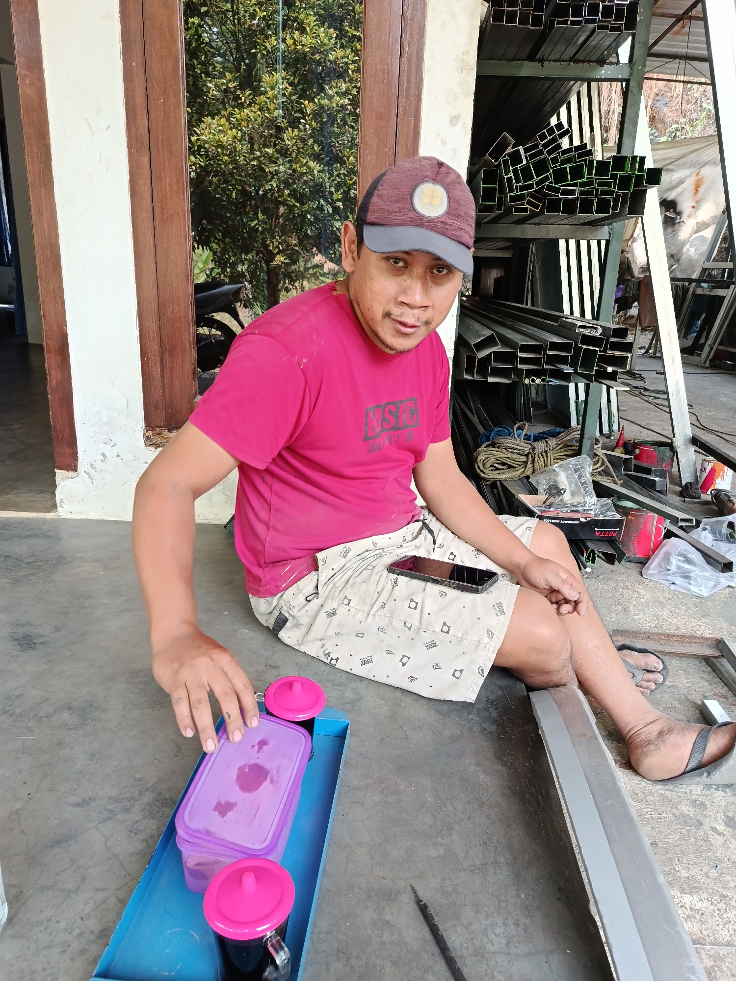A man wearing a red t-shirt and patterned shorts sits on a concrete floor in front of a building. He is wearing a baseball cap and sandals. A blue tray with two cups of coffee and a purple container is in front of him. He is reaching for the container with his left hand. Behind him, metal pipes are stacked on shelves. The scene appears to be a workshop or storage area. The location is in Lempong, Indonesia.