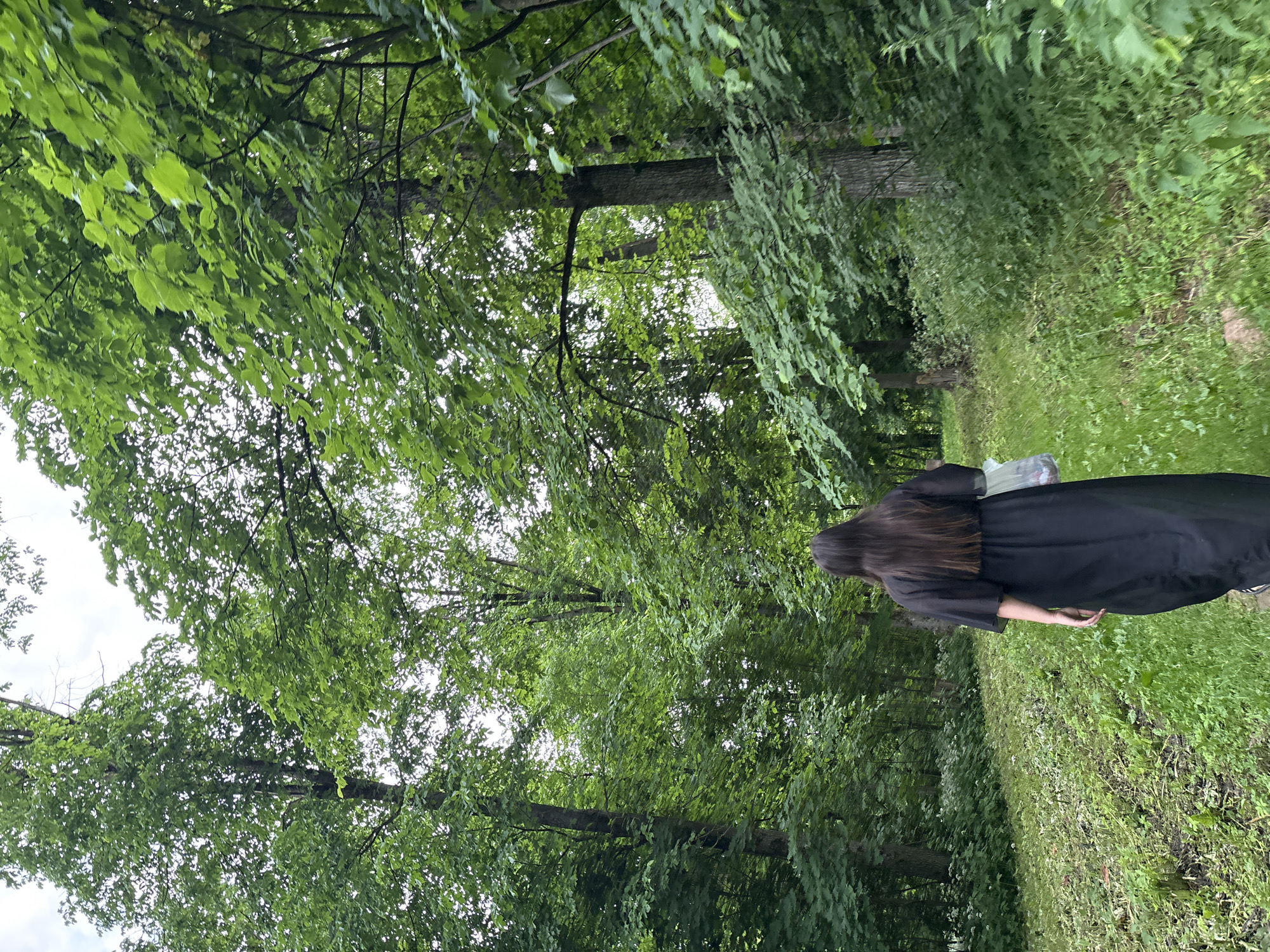 A person with long, dark hair, wearing a flowing black dress, walks away from the camera on a path through a lush green forest. The path is covered in grass and some scattered leaves. Tall trees with dense green foliage surround the path, and the sky is visible through the branches. The person is carrying a clear plastic bag in their right hand. The scene appears to be outdoors, likely in a park or wooded area within the city of Gorki, Russia.