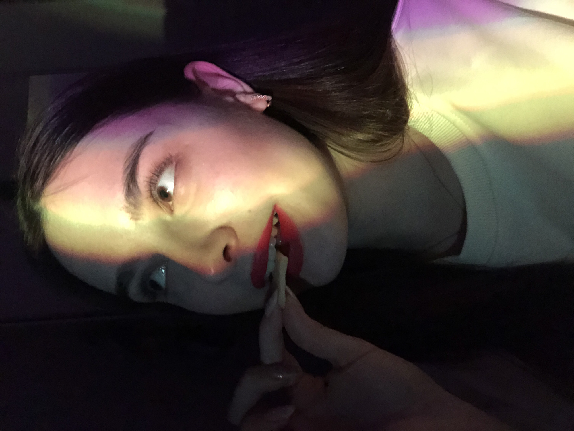 An individual with long, dark hair and fair skin is depicted lying down, oriented horizontally in the frame. Their face is illuminated by a striking array of projected colored lights, casting vibrant purple, pink, yellow, and green hues across their features and hair. The background is dark, suggesting a dimly lit indoor setting, likely at night or evening.
The person is holding a rolled, light-colored object, consistent with a cigarette or joint, between their red-lipsticked lips and the fingers of one hand. Their eyes are wide open, gazing upwards and to the right with an intense or pensive expression. A small earring is visible on their ear. Part of a light-colored, ribbed garment, possibly a sweater or sweatshirt, is visible near their shoulder. No text is visible in the image.