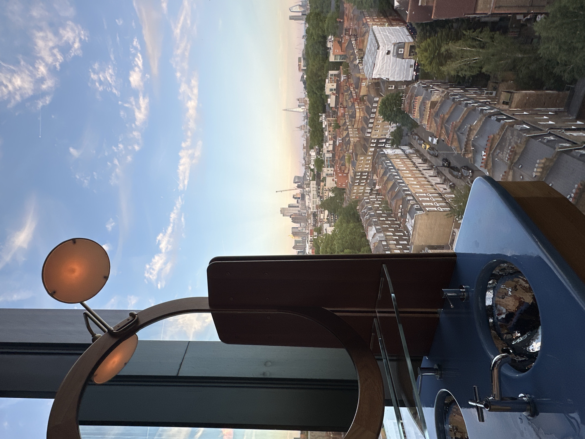 From a high vantage point, possibly inside a building, a view of London unfolds under a sky with scattered clouds. The urban landscape features densely packed buildings with rooftops visible, interspersed with green spaces from trees. Landmarks punctuate the skyline, including the Shard, a tall, pyramidal skyscraper.
Foreground elements include interior details: a light fixture with round shades, and a blue washbasin with metallic fixtures. The combination suggests an indoor setting overlooking the cityscape.