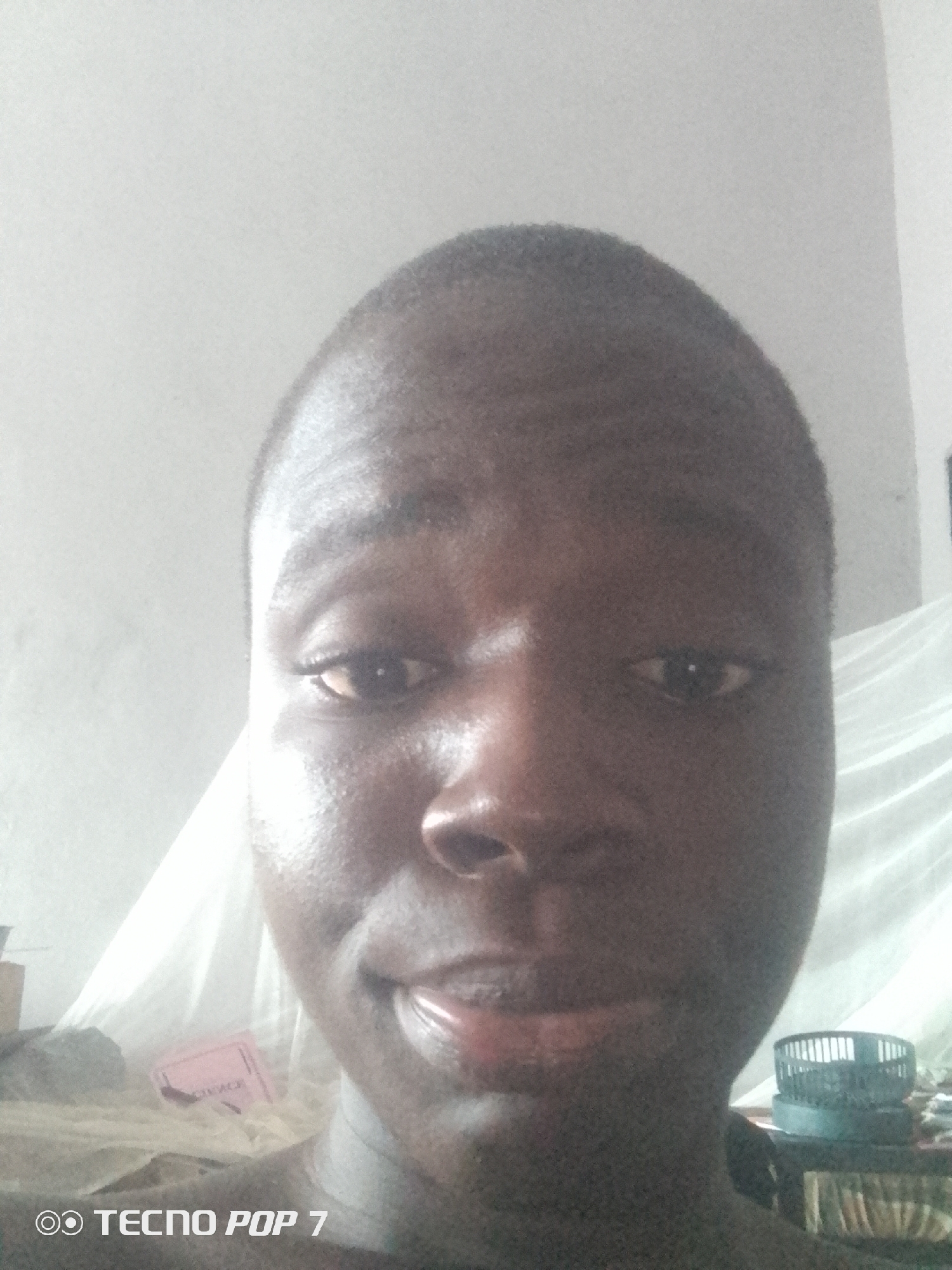 A close-up, selfie-style portrait of a young Black male is captured from the chest up. The subject has short, dark hair and is looking directly at the camera. His skin appears dark and is slightly shiny, possibly due to lighting or sweat. The background is out of focus but suggests an indoor setting with a white or light-colored wall and a draped fabric. To the left and slightly behind the subject, a pink object with some text is visible. To the right, a small, dark fan with a cage-like grille sits on a surface. The bottom left corner of the image displays a watermark that reads "TECNO POP 7". The image was taken in Ipetumodu, Nigeria.