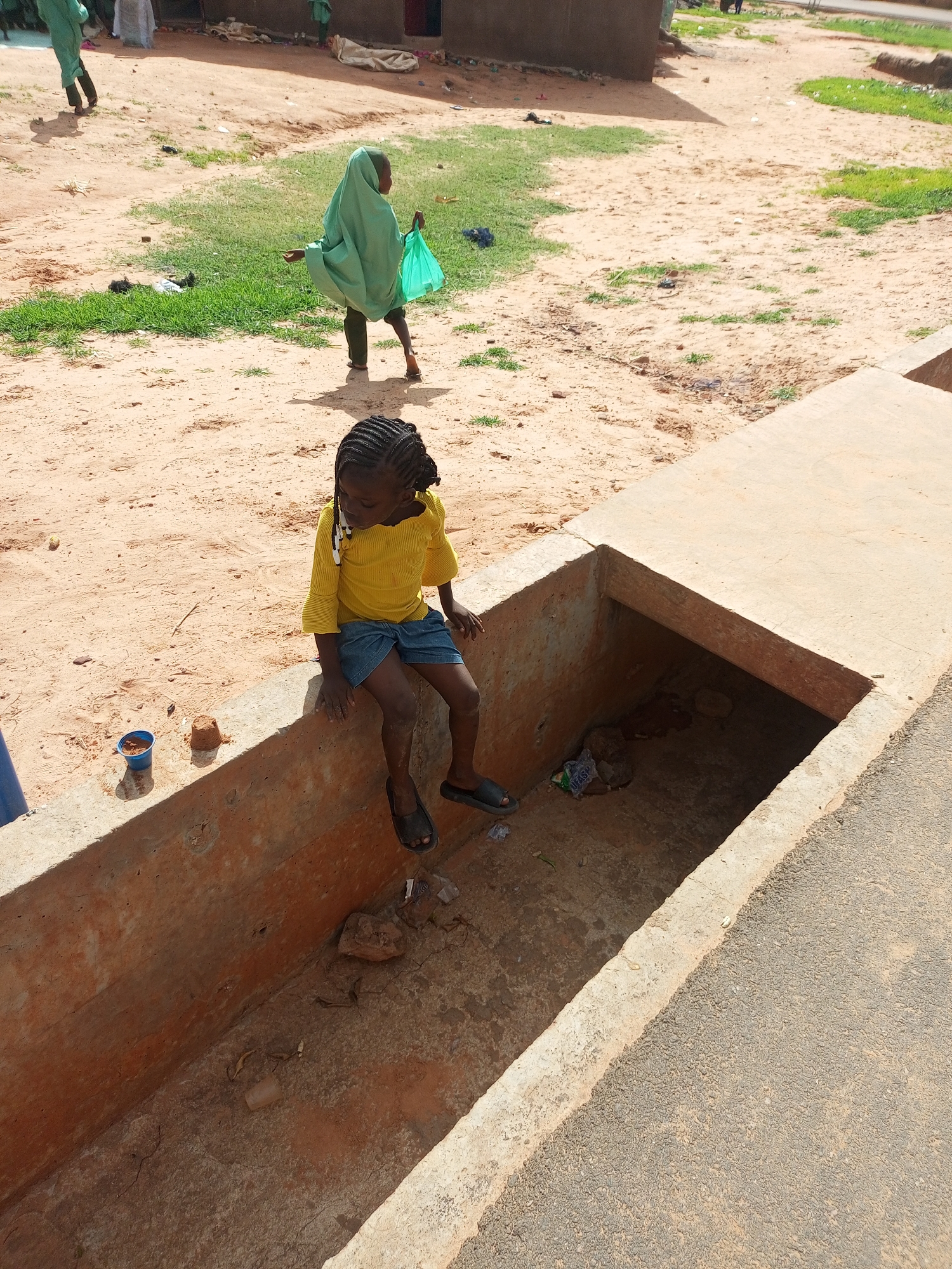 In a dusty, outdoor environment near buildings in Tempure, Nigeria, a young girl with braided hair sits on the edge of a concrete drainage ditch. She wears a yellow, ruffled-sleeve shirt and a blue skirt. Her feet are bare, clad in dark sandals. The drainage ditch is partially filled with dirt, rocks, and debris, including plastic bags. To her left, near the edge of the ditch, are a small blue cup and a mound of dirt. In the background, another child in a green hijab walks away, carrying a green plastic bag. Other figures and structures are visible in the distance, set against a backdrop of dry, sandy ground with patches of green grass.