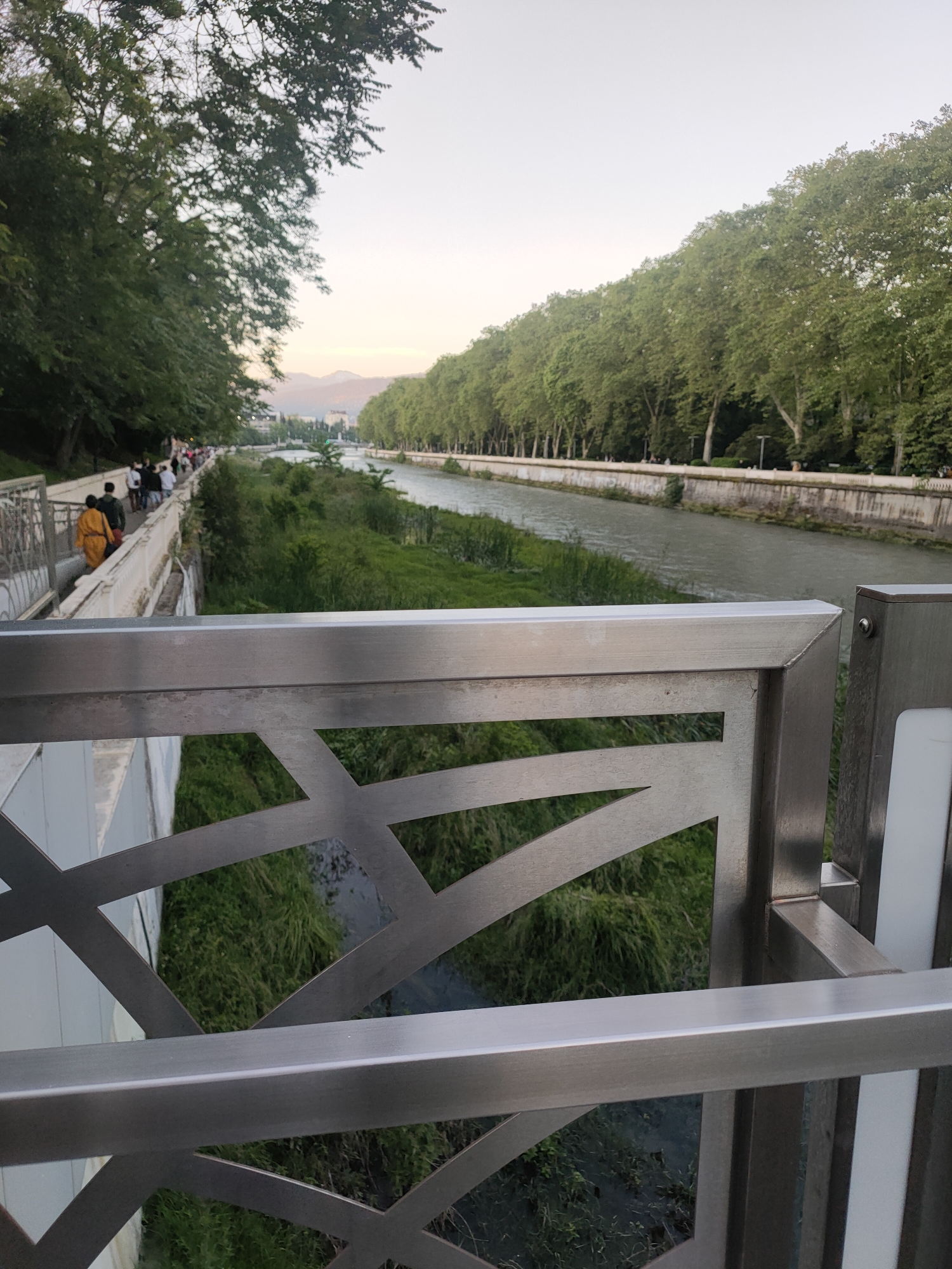 This photograph presents a scenic view of a tree-lined river or canal in Sochi, Russia, captured from an elevated position, likely a bridge or an overpass. The scene suggests late afternoon or early evening, characterized by soft, diffused light and a pale, almost golden sky above the distant mountains.
In the immediate foreground, a modern, brushed metal railing with a distinctive geometric pattern of angular cut-outs spans the lower part of the frame. A white light fixture is integrated into the railing structure on the right side. Beyond this railing, a calm body of water flows, flanked by dense rows of lush, green deciduous trees on both sides, creating a verdant corridor.
On the left bank, a paved promenade runs alongside the river, where several people are seen walking leisurely. One person in a vibrant yellow or orange top stands out among the group. The area between the pathway and the water is covered with green grass and foliage. The right bank also features a continuous line of tall trees. In the far background, hazy outlines of mountains are visible, lending a picturesque backdrop to the urban-natural landscape. The setting conveys a sense of tranquility and a popular recreational area. No discernible text is visible in the image.