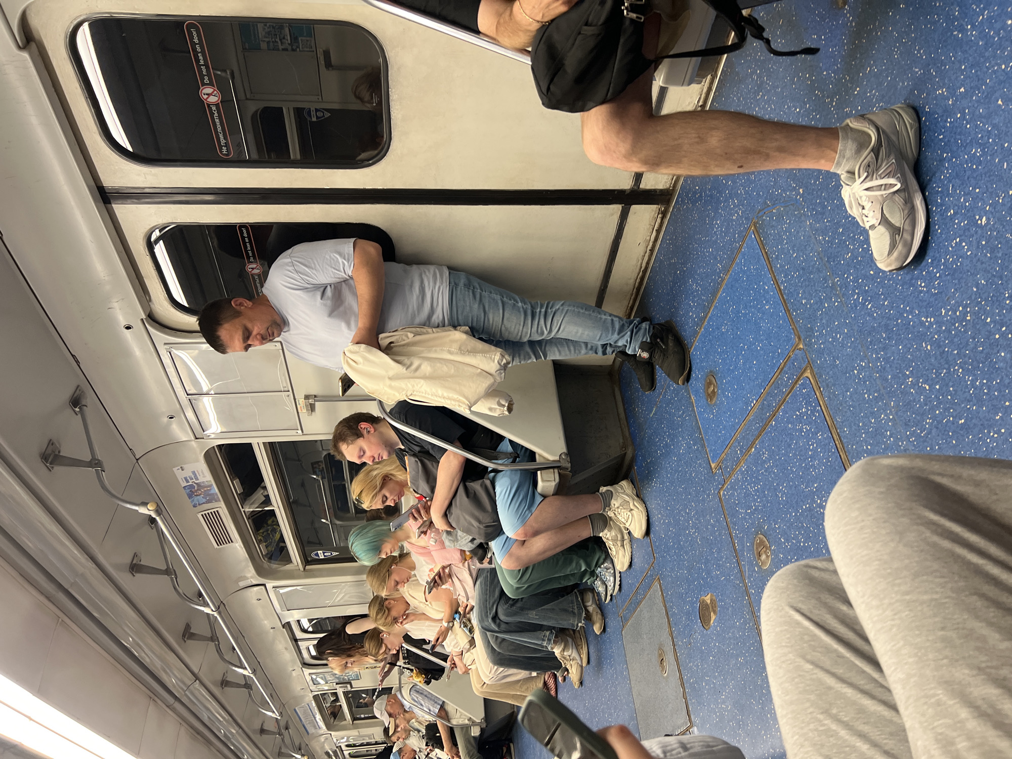 The image shows the interior of a subway car, with passengers seated and standing. The car has blue flooring with white speckles, and a white and gray interior. A man in a light blue t-shirt and jeans is leaning against a door, holding a cream-colored jacket. Other passengers are visible, some looking at their phones, others appearing to rest. One person in the foreground has their legs and feet visible, wearing grey socks and white sneakers. Visible text on a door reads "Do not lean on door!" and "Не прислоняться!". The lighting suggests it is daytime. The overall scene depicts a typical commute on public transportation.