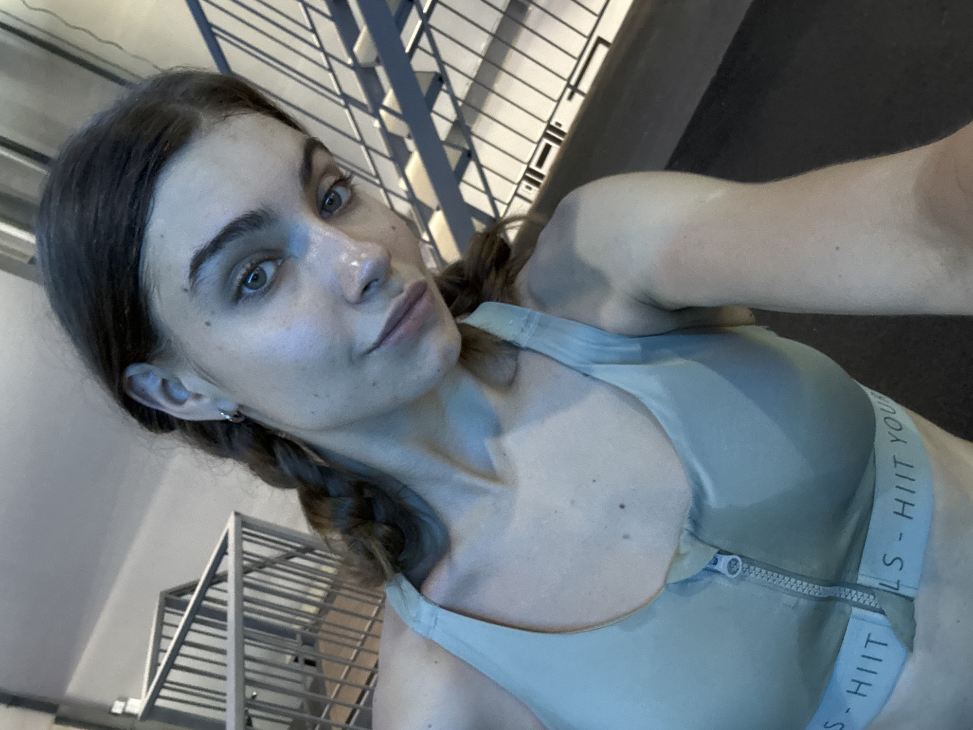 A woman with light skin, dark hair styled in two braids, and blue eyes takes a selfie indoors. She is wearing a light green sports bra with the words "LS - HIIT YOU" printed on the straps. The background includes a gray metal structure, possibly stairs or a rack. The location is not identifiable as Đà Nẵng, Vietnam.
