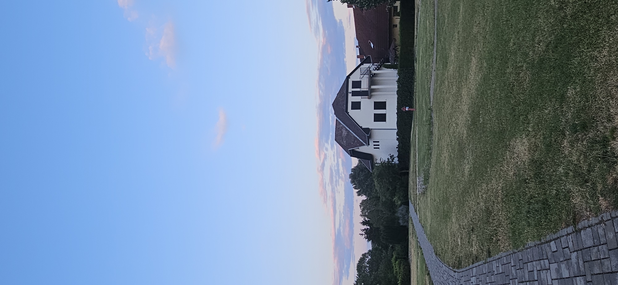 The image depicts a suburban scene in Hévíz, Hungary, under a twilight sky. A two-story white house with a dark roof and a balcony is the focal point, situated behind a manicured hedge. The house has several dark-paned windows and an external staircase leading to the upper floor balcony. In front of the house, a red fire hydrant is visible.
To the left of the house, a winding paved walkway made of gray interlocking bricks curves through a grassy area. The grass appears dry in some patches, suggesting a summer or late summer setting. Behind the house and walkway, a dense line of green trees forms the backdrop, with a glimpse of another house with a reddish-brown tiled roof to the right.
The sky above is a clear blue, transitioning to lighter shades near the horizon, with wisps of pink and white clouds scattered across it, indicative of sunset or sunrise. The overall impression is one of a quiet, residential neighborhood.