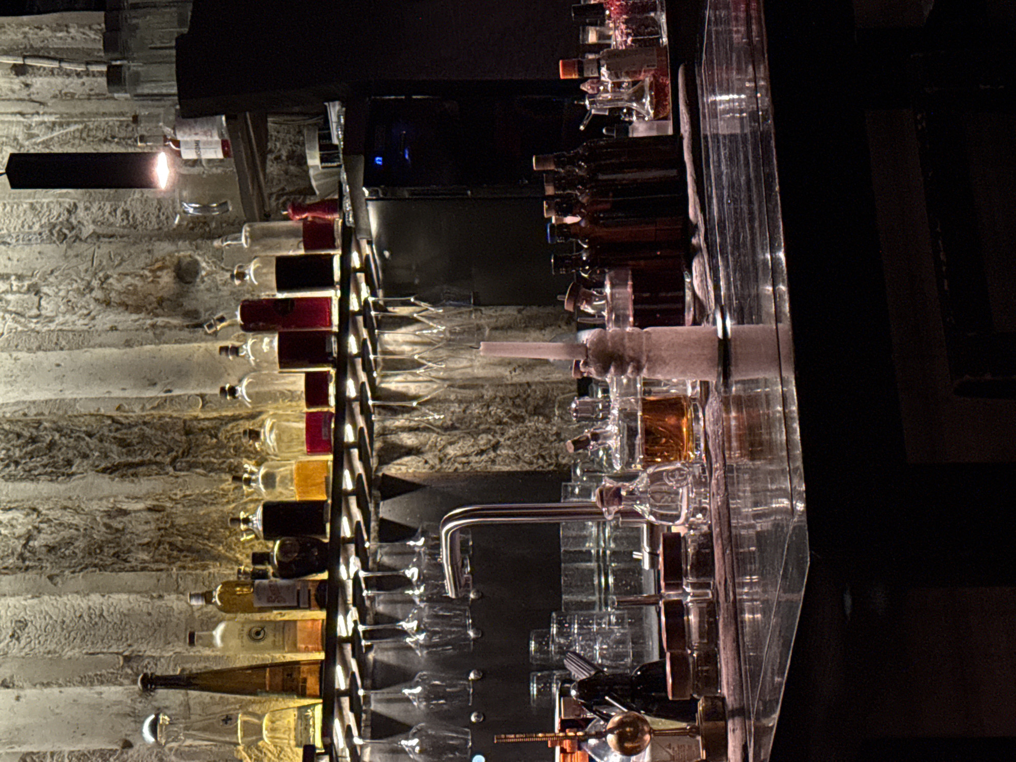 The image depicts the interior of a bar, likely in Moscow, Russia. The background features a stone wall with recessed shelves holding a variety of liquor bottles, illuminated by a linear light fixture above. In the foreground, a clear bar surface reflects rows of glassware and bar tools. A stainless steel faucet is also visible, suggesting a serving area. The overall scene is dimly lit, creating a sophisticated, potentially upscale, atmosphere.