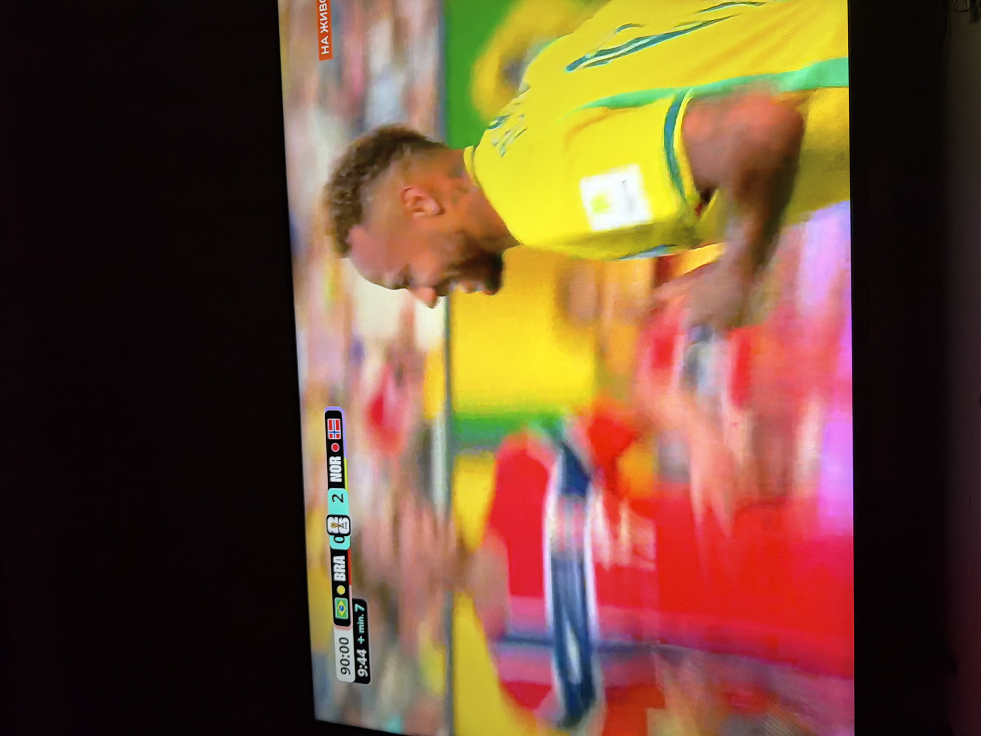 The image is a television screen displaying a football match with a score overlay. On the left side of the overlay, it shows "90:00", the Brazilian flag, "BRA", a score of "0", a Norwegian flag, and "NOR" with a score of "2". Below this, it reads "9:44 + min. 7". On the right side of the screen, a man in a yellow jersey with green trim is visible in profile, smiling. He appears to be a football player. Another player in a red jersey with blue and white stripes is blurred on the left. The background is out of focus, showing stadium lights and blurred spectators. A red banner with white Cyrillic text, "НА ЖИВО", is visible in the top right corner of the screen, indicating "LIVE" broadcast. This scene is a television broadcast and not a real-world location in Ognianovo, Bulgaria.