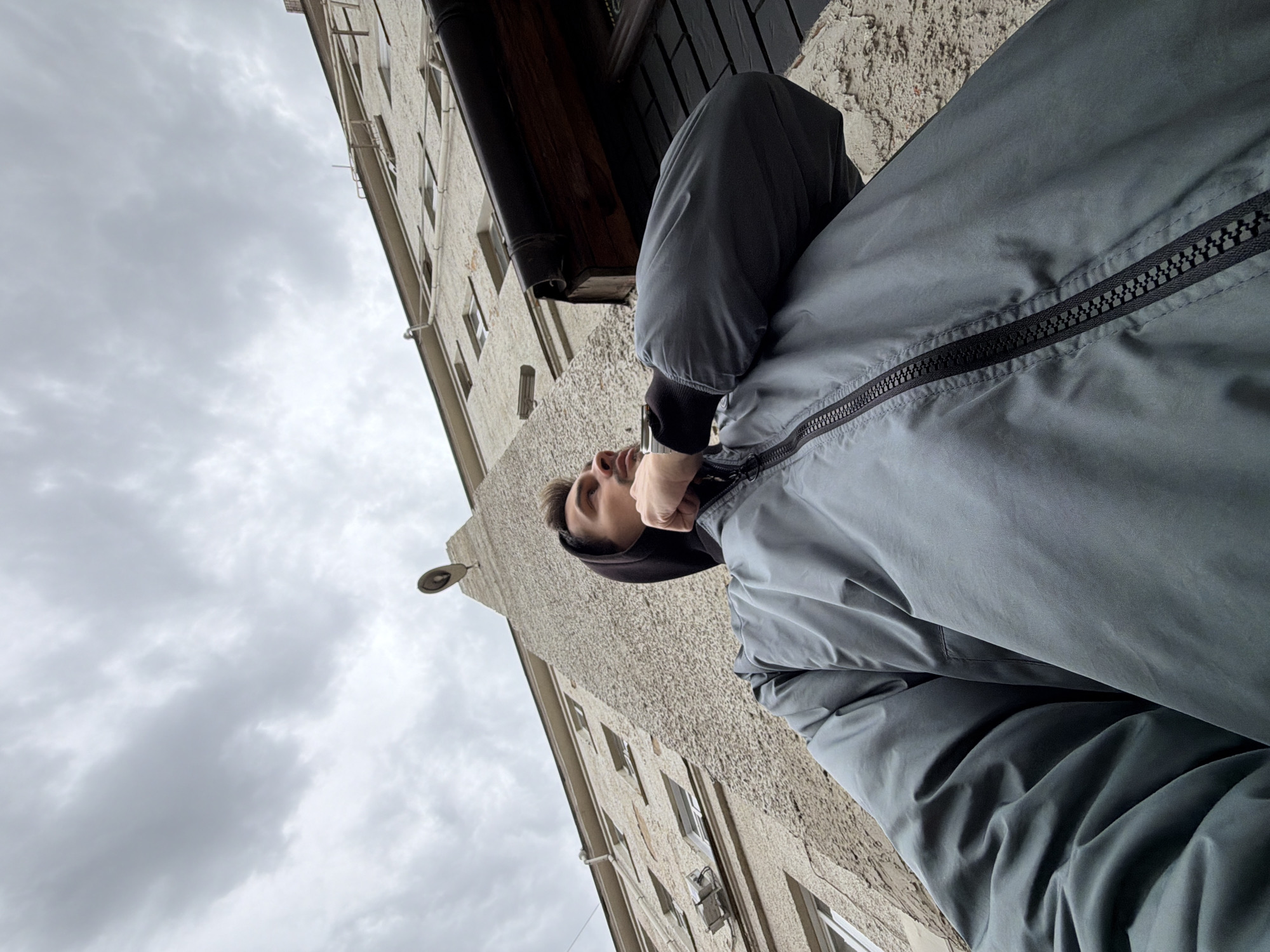 A low-angle shot captures a person in a gray hooded jacket, looking up at a building. The building is a pale concrete structure with multiple windows. A dark wooden element protrudes from the building, possibly a window frame. A streetlight fixture is visible on the building's exterior. The sky is overcast. The location is Krasnoyarsk, Russia.