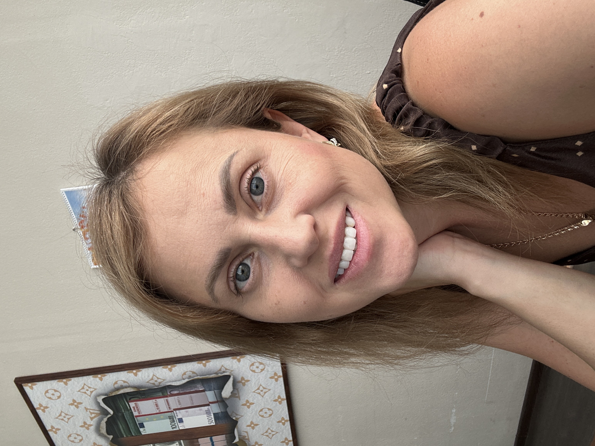 A selfie of a Caucasian woman with blonde hair and blue eyes, smiling and resting her chin on her hand. She is wearing a brown dress with a small white polka dot pattern and a gold necklace. In the background, there is a framed picture with a Louis Vuitton-inspired pattern and images of euro banknotes. The wall behind her is a light beige color. The scene appears to be indoors, likely in a home or office setting. No specific landmarks or indicators of Sochi, Russia are visible in the image.
