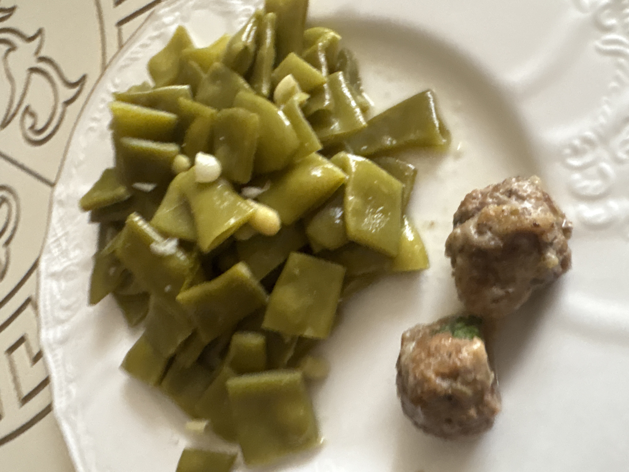 A close-up overhead shot shows a white ceramic plate with a decorative rim, filled with cooked green beans and two small meatballs. The green beans are cut into flat, diamond-like shapes and appear to be stewed in a light sauce with visible pieces of garlic. The meatballs are round and have a browned exterior, suggesting they are cooked. The plate rests on a patterned tablecloth. The image does not contain any information about the city of Georgiyevsk, Russia, nor does it depict any people or actions.