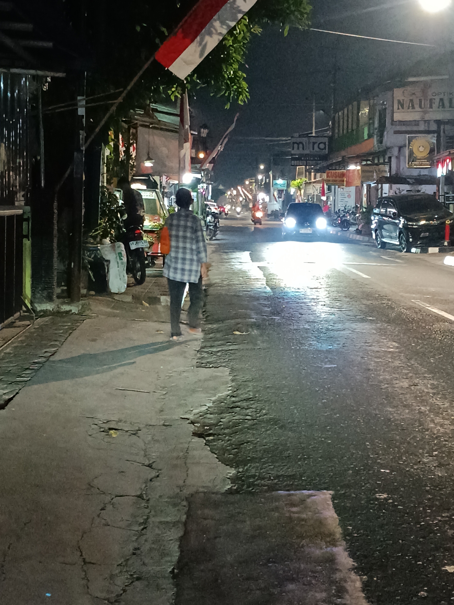 A person walks on a sidewalk at night on a street in the city of Yogyakarta, Indonesia. The person is wearing a plaid shirt and dark pants. The sidewalk is made of concrete and has cracks. The street is wet and reflective from streetlights. There are cars and motorcycles on the street. Buildings are visible on the right side of the street. A flag, possibly the Indonesian flag, is hanging from a pole on the left side of the street. The overall scene is typical of a street in a city at night, with activity and lighting from businesses and vehicles.