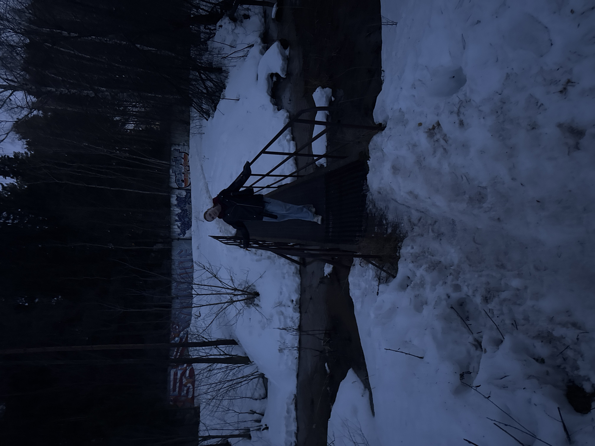 The image depicts a person standing on a metal grate bridge over a narrow stream or ditch. The location is snowy, suggesting it is winter or early spring. Trees are visible in the background. The individual is wearing a dark jacket and light-colored pants. Graffiti can be seen on a wall near the tree line, possibly indicating the location is urban or semi-urban. The scene seems dimly lit, possibly taken during twilight or in low light conditions. It is most likely located in Chaikovsky, Russia.