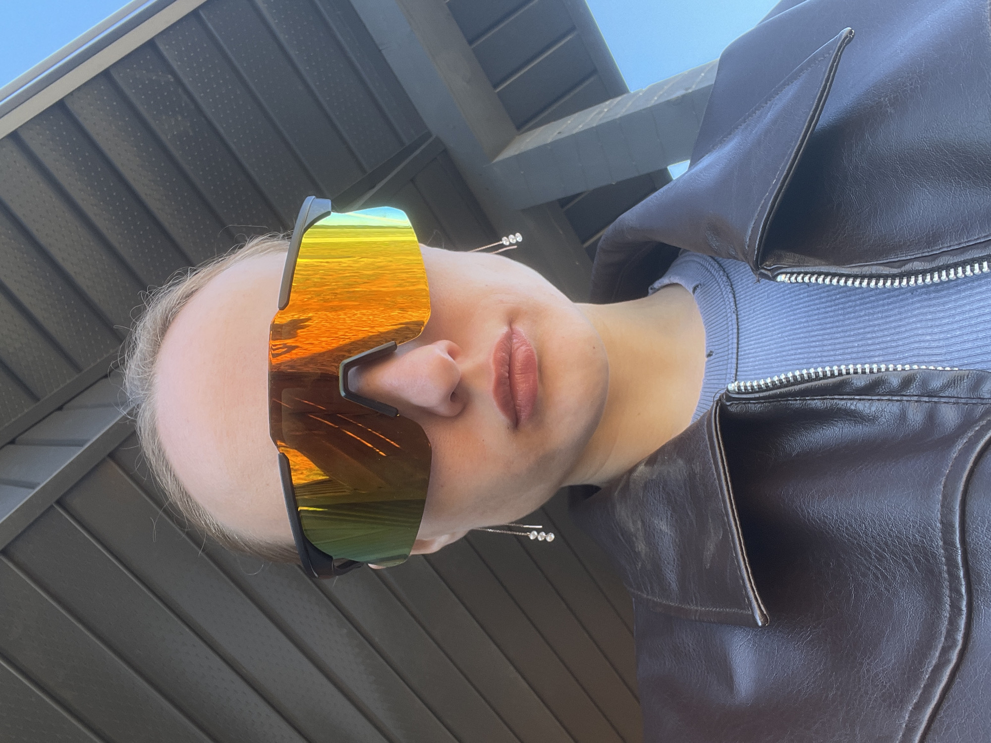 A person is seen from a low angle, wearing reflective sunglasses that obscure the eyes. The reflection in the sunglasses appears to be a horizon at sunset or sunrise. The person has fair skin and is wearing a light-colored top underneath a dark brown leather jacket. The jacket is partially zipped. The person is wearing an earring consisting of a thin metal bar with three small, possibly gem-set, spheres. The background consists of an overhanging gray corrugated metal structure, above which a blue sky is visible. There is no information available in the image to indicate that the location is in Подшивалово, Russia.