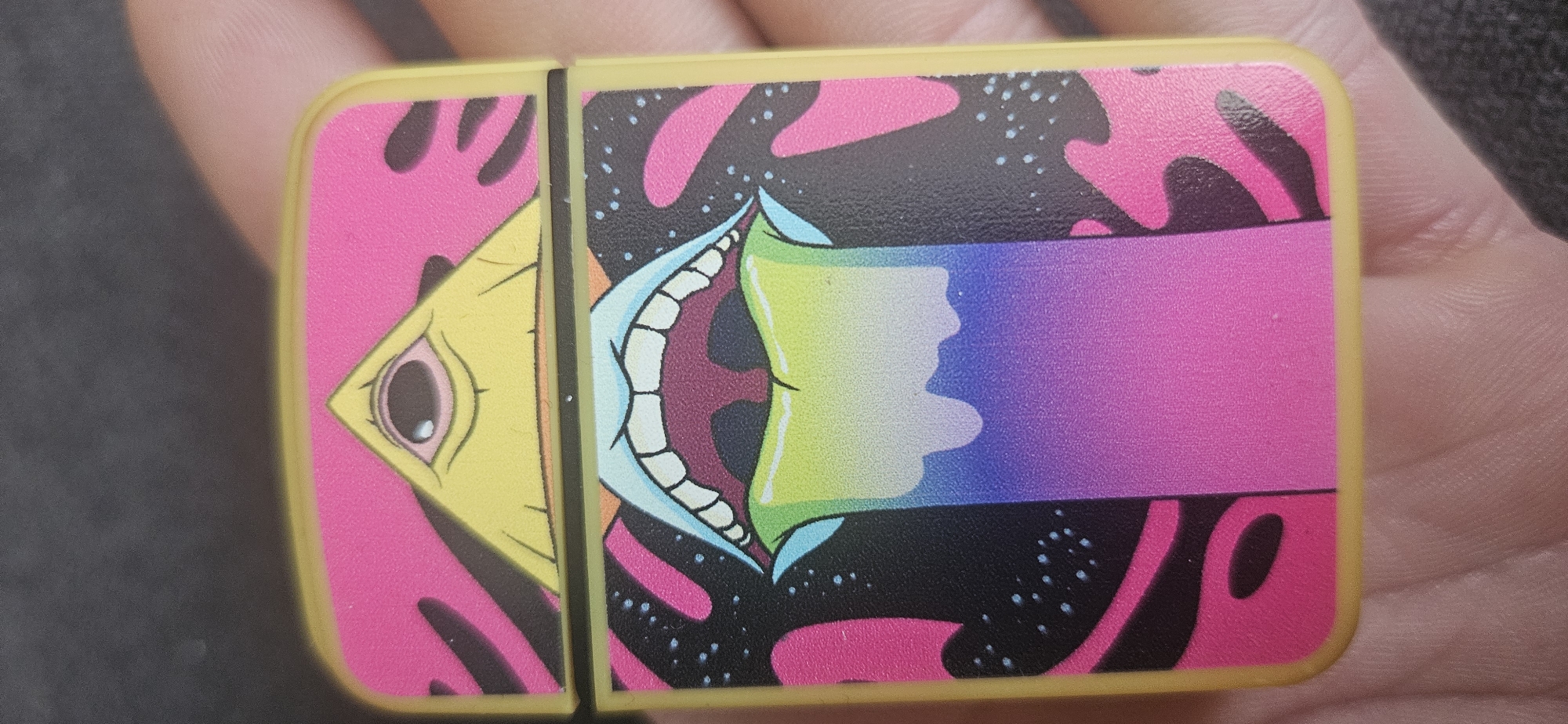 A close-up shows a yellow rectangular object, likely a container, held in a hand. The container has an illustration featuring a yellow, triangular face with a large eye on the left side. To the right, there is a large, open mouth with teeth. From the mouth emanates a gradient of colors, transitioning from green to purple. The background includes pink and black organic shapes with white specks. The object has a yellow border. No information indicates the location of Plovdiv, Bulgaria.