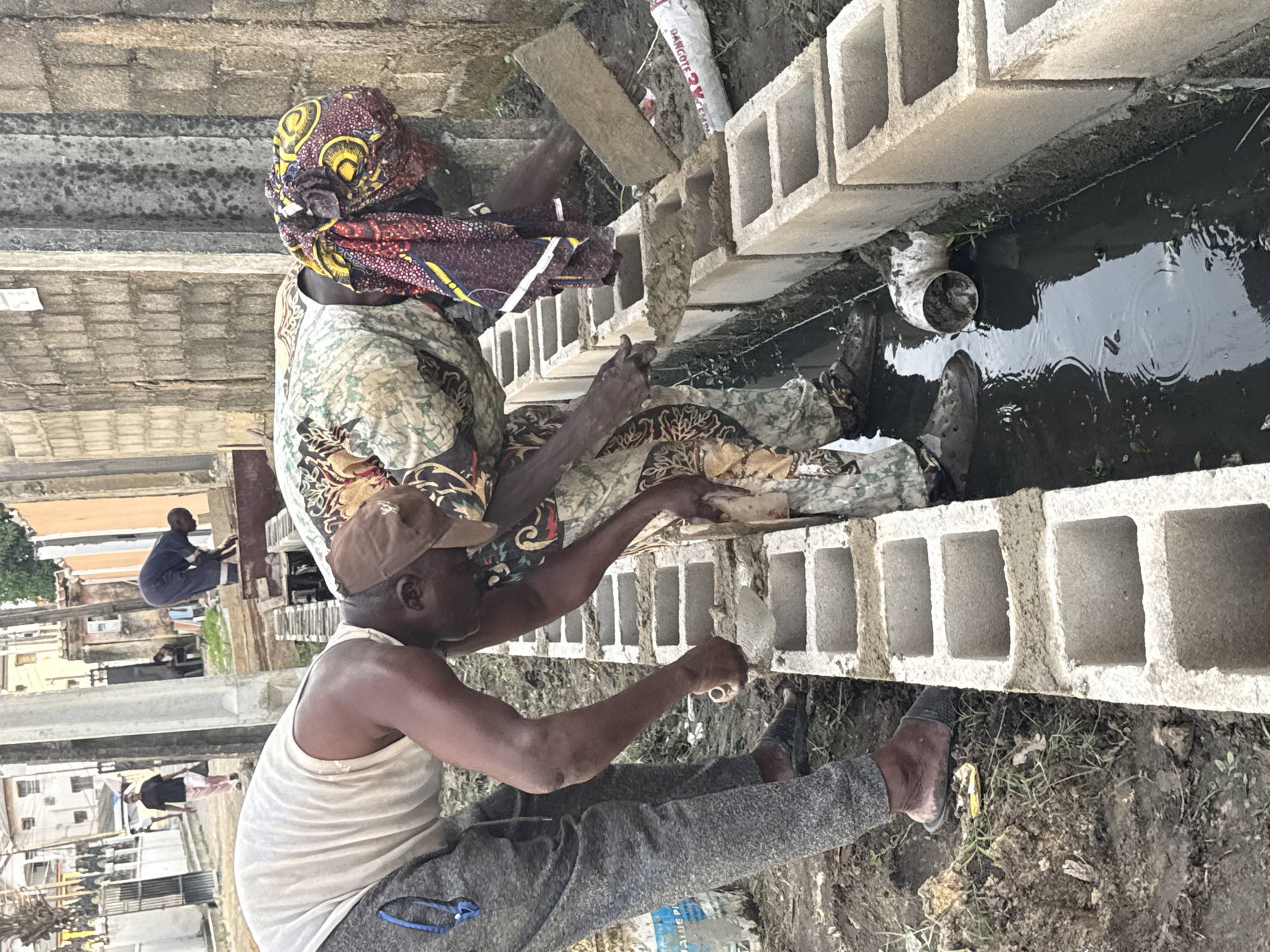 Two individuals are laying concrete blocks along a narrow, water-filled ditch. The person closer to the camera is wearing a tan baseball cap, white tank top, and gray sweatpants, and they are using a trowel to apply mortar. The second person, sitting behind them, is wearing a patterned head covering and clothing. The concrete blocks are gray and have rectangular openings. The ditch is filled with dark water, and a white PVC pipe is visible in the water. In the background, there are buildings and other individuals visible, suggesting an urban environment. The location is Ado, Nigeria.