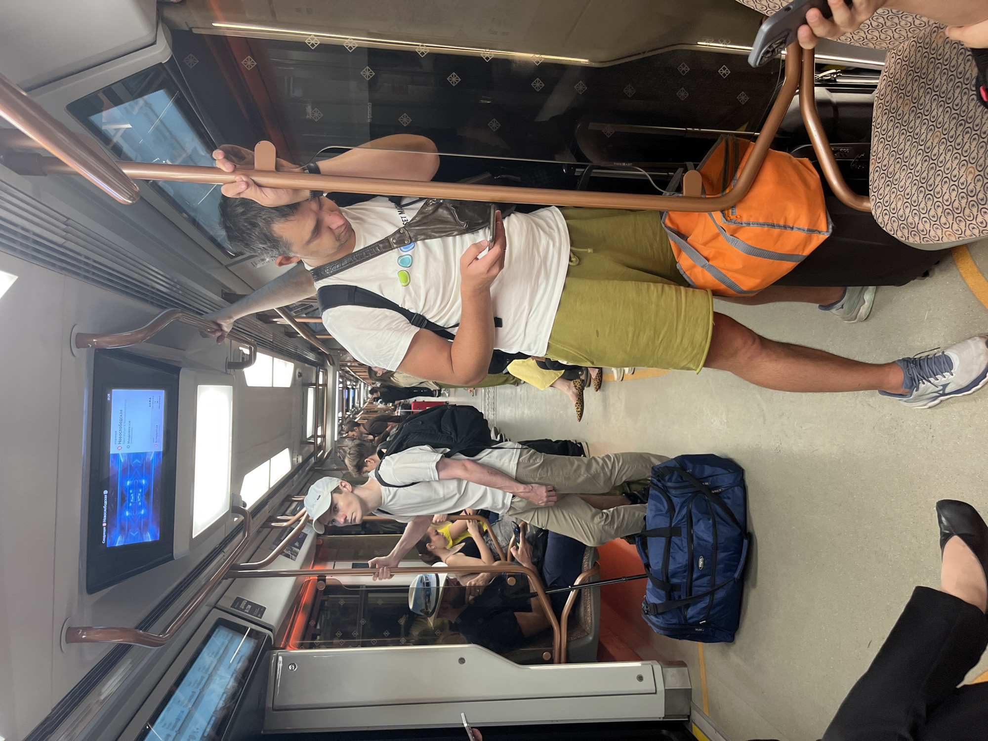 The image depicts the interior of a Moscow Metro train. Several passengers are visible. In the foreground, a man wearing a white t-shirt and olive-green shorts is holding a smartphone and leaning on a handrail. He has a brown crossbody bag and a black backpack. Next to him, a large blue duffel bag rests on the floor. Further down the car, another man in a white t-shirt and khaki pants stands holding a handrail, wearing a white baseball cap and a black backpack. Other passengers are seated and standing throughout the train car. A digital display above shows station information for "Новослободская" (Novoslobodskaya) and "Менделеевская" (Mendeleevskaya), confirming the location within the Moscow Metro system. The train car features brown handrails and decorative patterns on the walls and seats. An orange bag is visible near the man in the foreground, placed on top of a dark suitcase. The lighting suggests it is daytime.