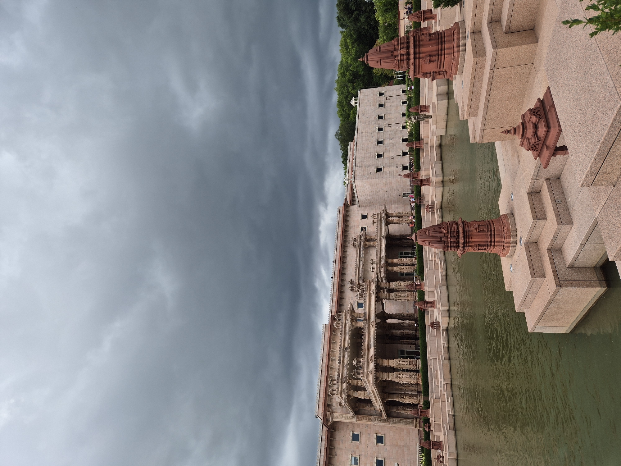 The image captures the exterior of a grand stone building, likely a temple or cultural center, in Robbinsville Township, United States of America. The architecture is ornate, with multiple levels, intricate carvings on pillars and facades, and a covered walkway supported by columns. In the foreground, a body of greenish water, possibly a reflecting pool or pond, is visible, with tiered stone platforms and decorative red spires emerging from the water and the surrounding stone structures. The sky above is heavily overcast with dark, dramatic clouds, suggesting an impending storm or recent rainfall. A few people are sparsely scattered in the background near the building entrance, and trees line the far edge of the property. The overall scene conveys a sense of solemnity and grandeur, amplified by the moody sky.