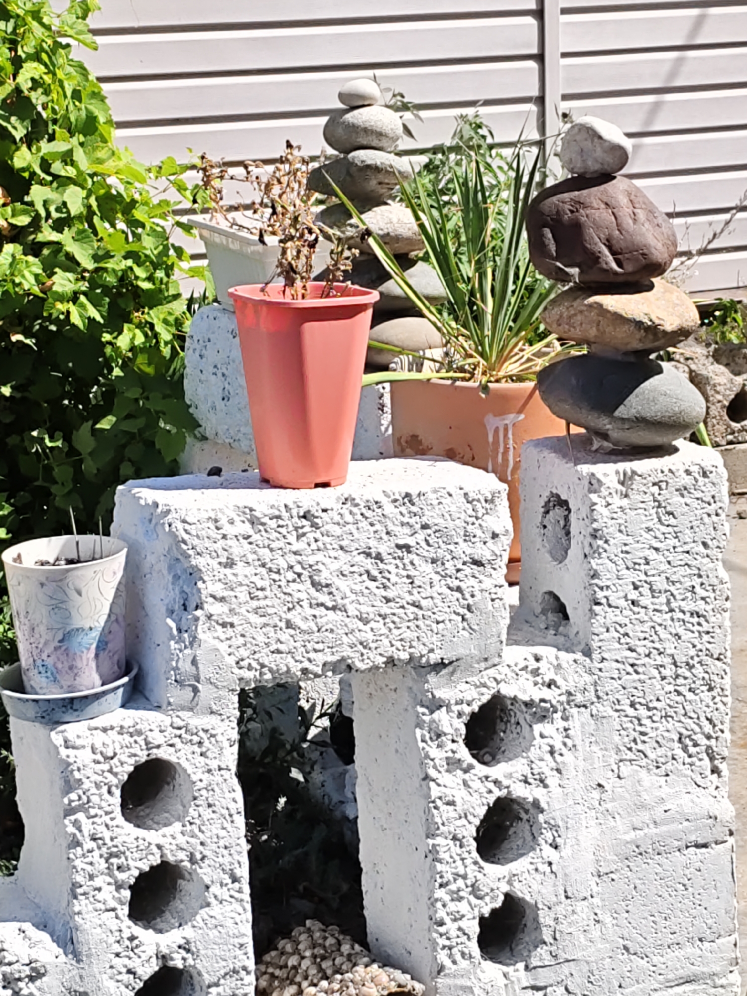 The image depicts a garden scene with a white, textured, cinder block structure in the foreground, painted with a rough, stucco-like finish. The structure has several openings and is topped with pots. One pot, a terracotta color, contains a dried plant. Another, a smaller white pot with a floral pattern, sits on a saucer. Behind the structure, a stack of smooth, grey and brown stones is balanced precariously. Green foliage and a tall, spiky plant are visible in the background, along with a light-colored, horizontal plank fence. The scene appears to be outdoors, under bright sunlight, suggesting a warm day. The setting is likely a residential garden in Rostov-on-Don, Russia, given the architectural style of the fence and the general appearance of the garden elements. There are no people or animals visible in the image.