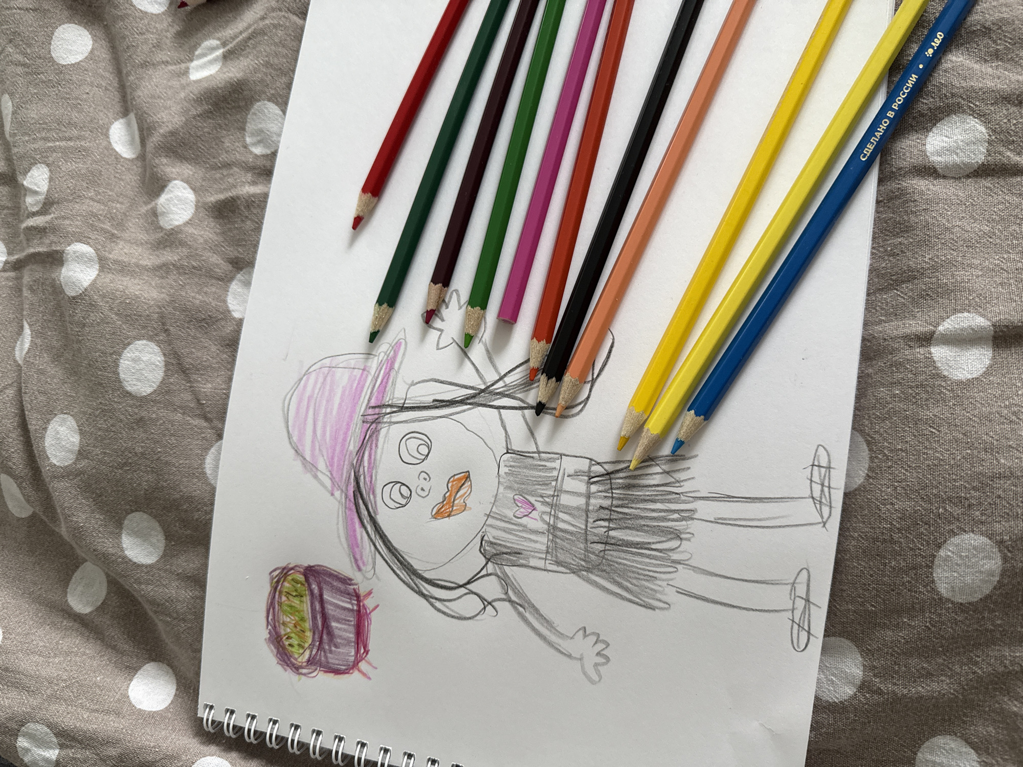 The image shows a sketch of a child's drawing on a white notepad. The drawing depicts a figure with a large head wearing a pink hat, an orange mouth, and a dress. To the left of the figure is a drawing of an object colored in purple and green. Several colored pencils are arranged diagonally across the top of the notepad, including red, green, brown, pink, orange, black, yellow, and blue. The notepad rests on a gray fabric with white polka dots.
The blue pencil has Cyrillic text that says "Сделано в России" (Made in Russia) and the letters "ADO". Izhevsk is not directly specified in the photo, but if the pencil is of the type manufactured by ADO, it would have been produced in Izhevsk.