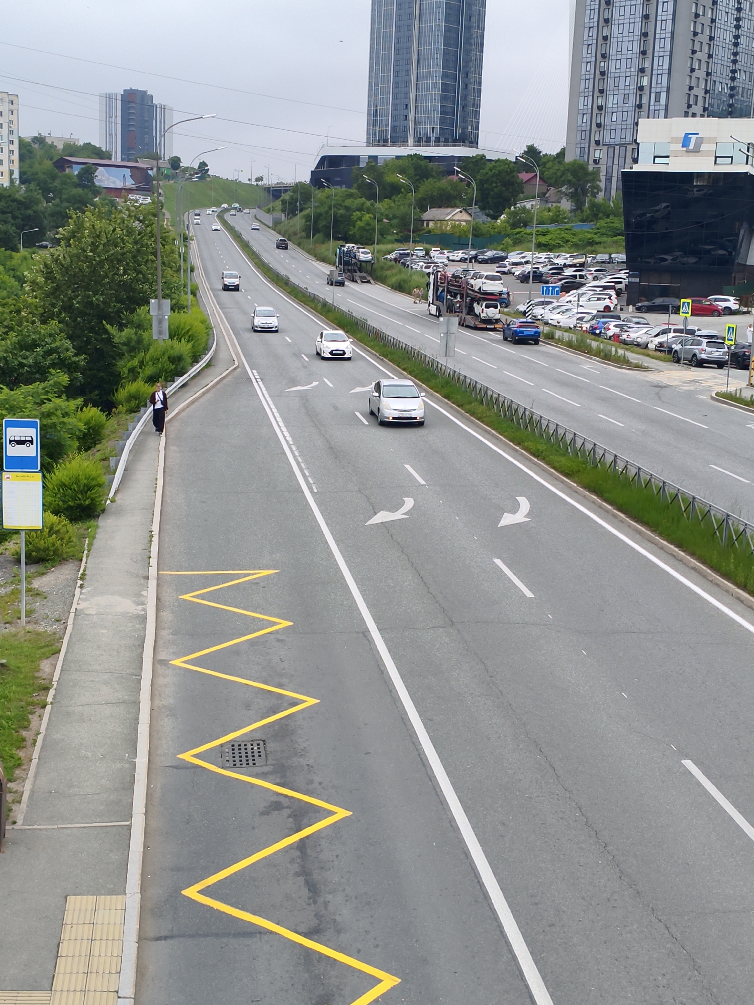This is a high-angle, medium shot of a multi-lane highway in Vladivostok, Russia, likely on a cloudy day. The highway curves to the left, with cars traveling in both directions. On the right side of the highway, there is a large parking lot filled with cars and a car carrier truck, in front of a modern glass-fronted building. On the left side of the highway, a woman in dark clothing is walking on a sidewalk next to a bus stop sign. There are also several tall buildings visible in the background, including one with a distinctive glass facade. The road is marked with white lines, and there is a yellow zigzag marking on the lane next to the sidewalk, indicating a no-parking zone. Green trees and bushes line the sides of the highway.