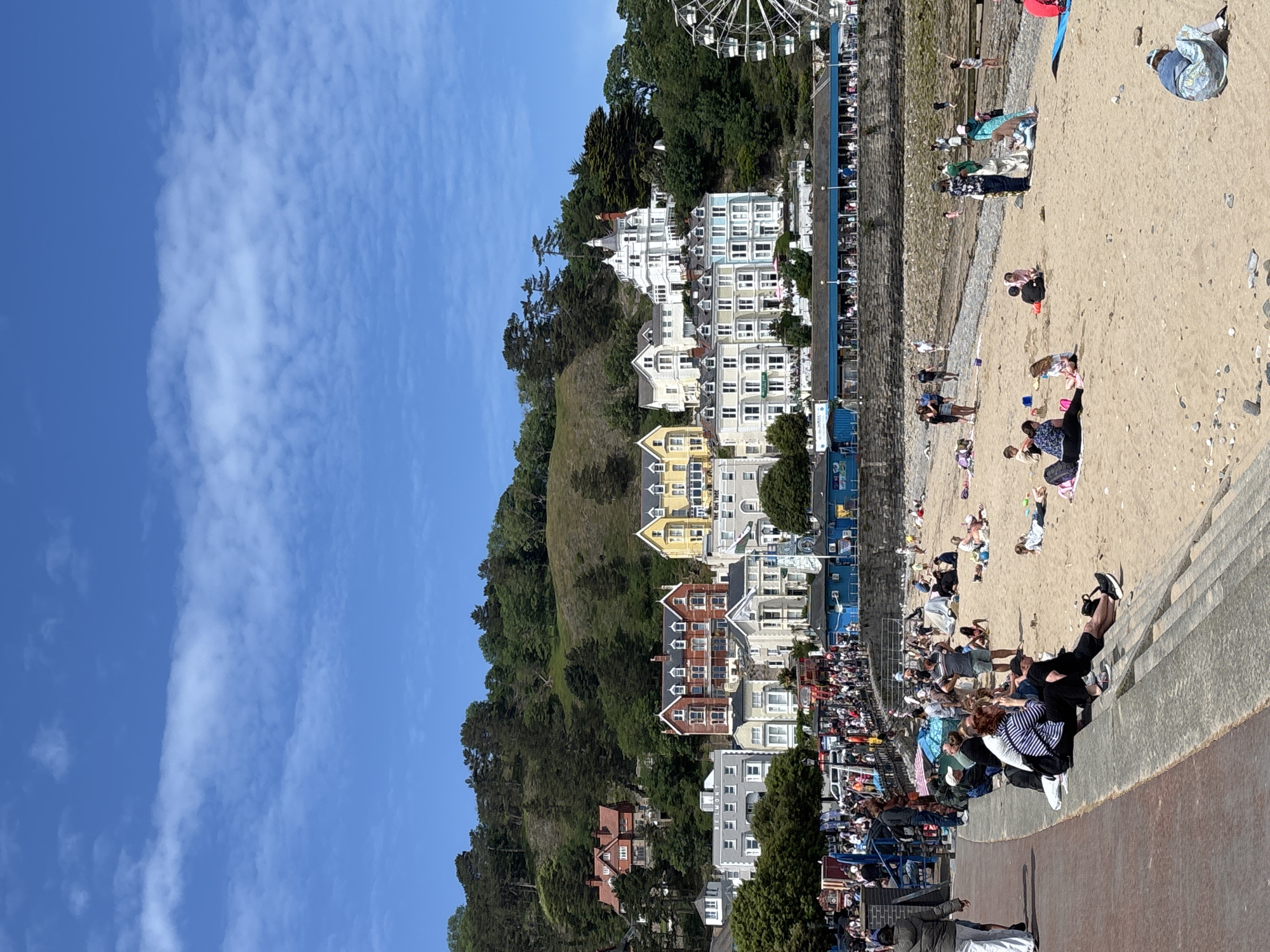 The image captures a vibrant scene at Llandudno Beach in the United Kingdom. The beach is crowded with people engaging in various activities, such as sunbathing, walking, and playing in the sand. Adjacent to the beach is a promenade lined with shops and seating areas, bustling with people. Behind the promenade, a steep hillside is densely populated with houses of varying colors and architectural styles. A ferris wheel is situated behind the houses. The sky above is blue with scattered clouds.