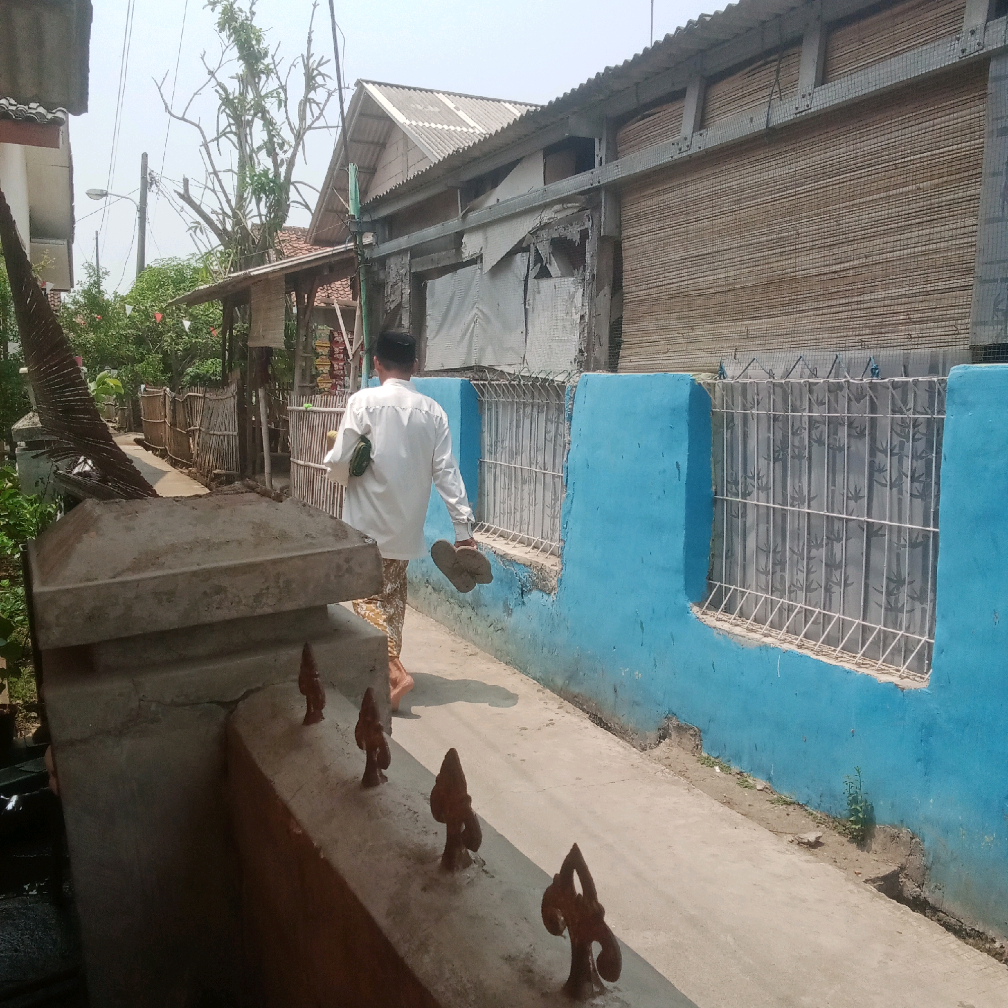 A man wearing a white long-sleeved shirt and patterned pants walks down a concrete path, carrying a pair of sandals in his left hand and a green object in his right. He is walking away from the camera towards a narrow street lined with buildings on the right and a bamboo fence on the left. The buildings appear to be made of corrugated metal and wood, with some showing signs of disrepair. The building on the right has a blue painted wall with a barred window. Vegetation and trees are visible in the background. The scene is set in Karawang, Indonesia.